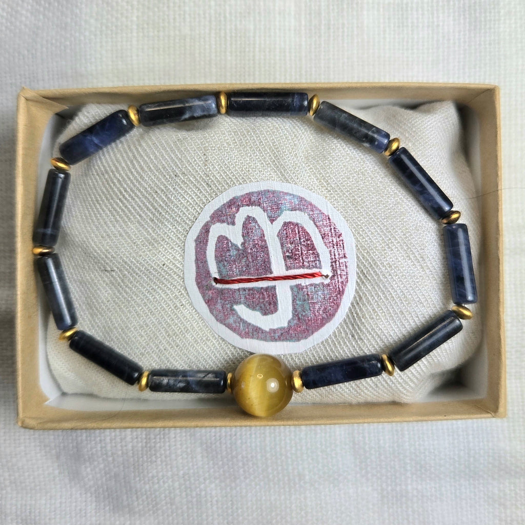 A beaded bracelet with dark blue-gray stone beads, small gold spacer rings, and a round gold tiger's eye bead, displayed inside a light beige gift box on a textured white fabric background.