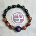 A beaded stretch bracelet arranged in a circle on a white textured fabric background, featuring dark black beads, brown glittery beads, green spacer beads, and a prominent purple-to-blue gradient focal bead. A circular sticker with a white symbol is placed behind the bracelet.