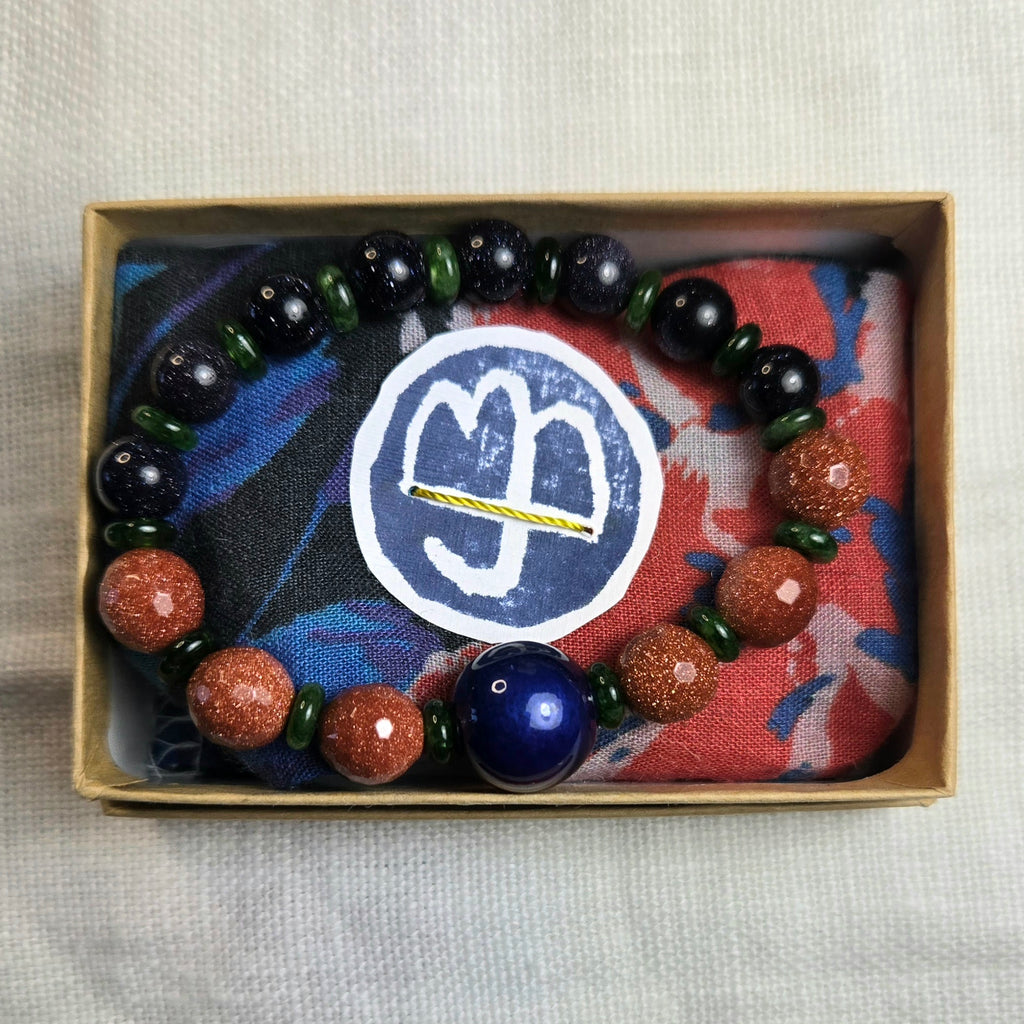 A beaded bracelet displayed inside a small kraft gift box on top of a colorful fabric insert. The bracelet consists of glossy black beads, translucent green beads, reddish-brown spiny oyster shell beads, and one large dark blue bead.