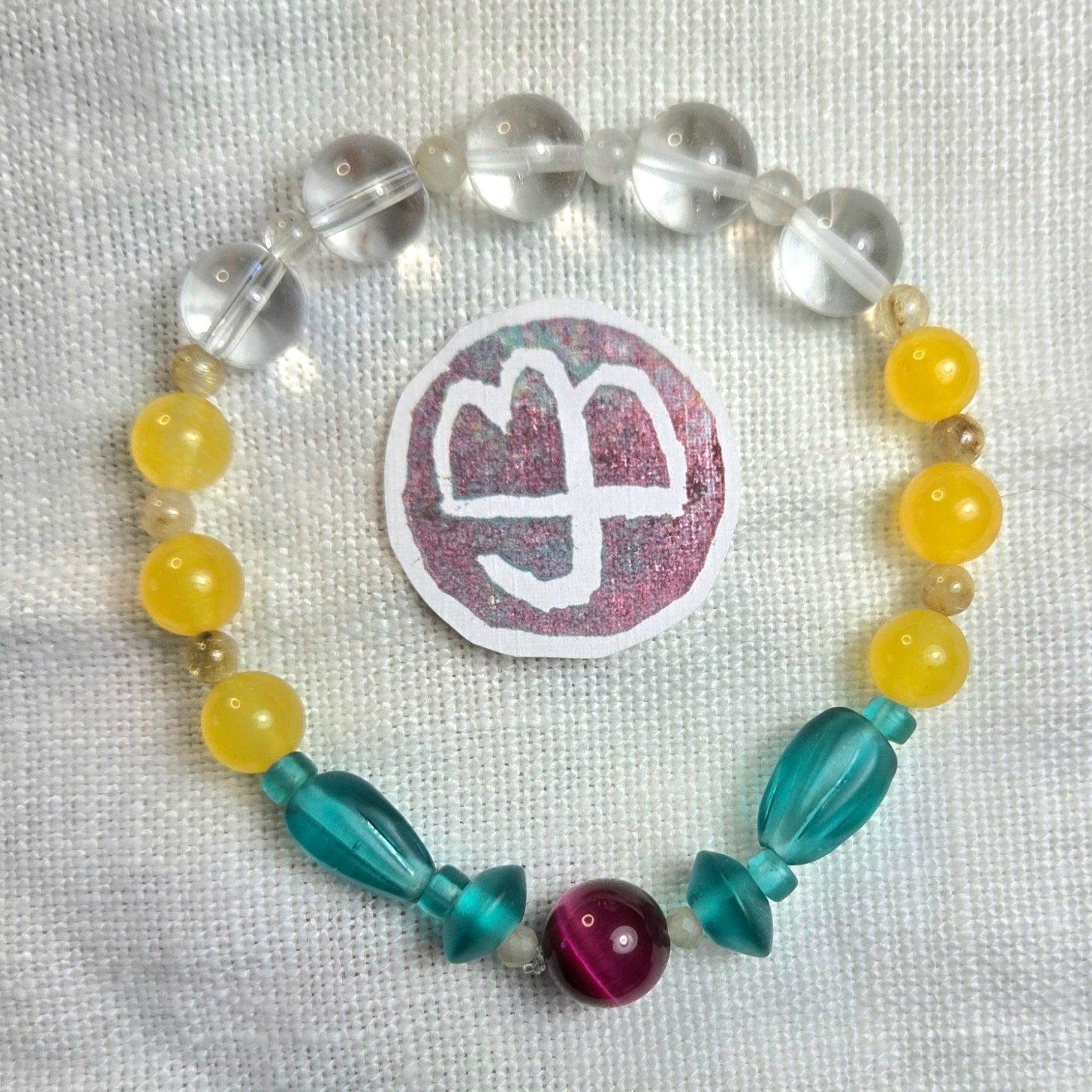 A beaded bracelet arranged in a circle on a textured white fabric background, featuring clear faceted beads, yellow round beads, teal translucent beads, a magenta bead, and a central glittery round patch with a white symbol. No brand is visible.