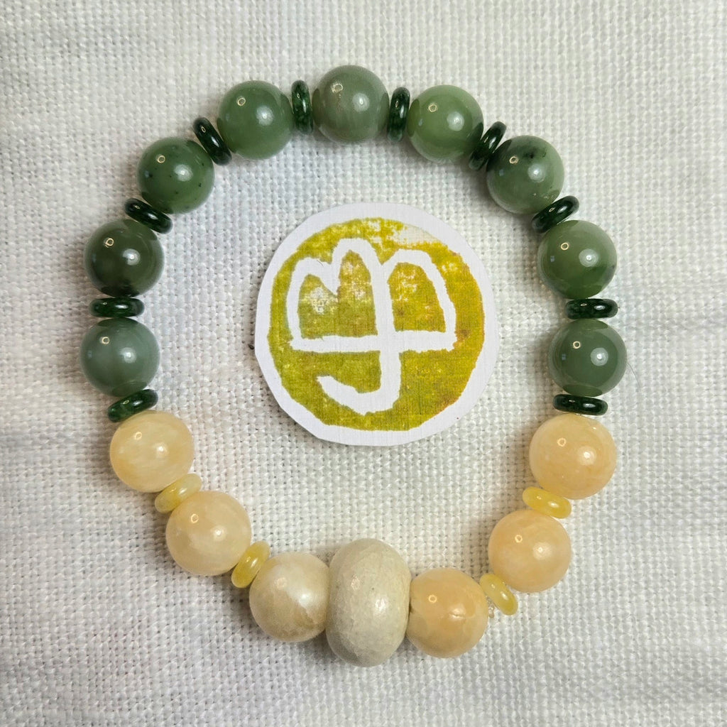 A beaded stretch bracelet with dark green marbled beads, pale yellow beads, and a central white circular charm featuring a gold-toned symbol, displayed on a textured white fabric background.