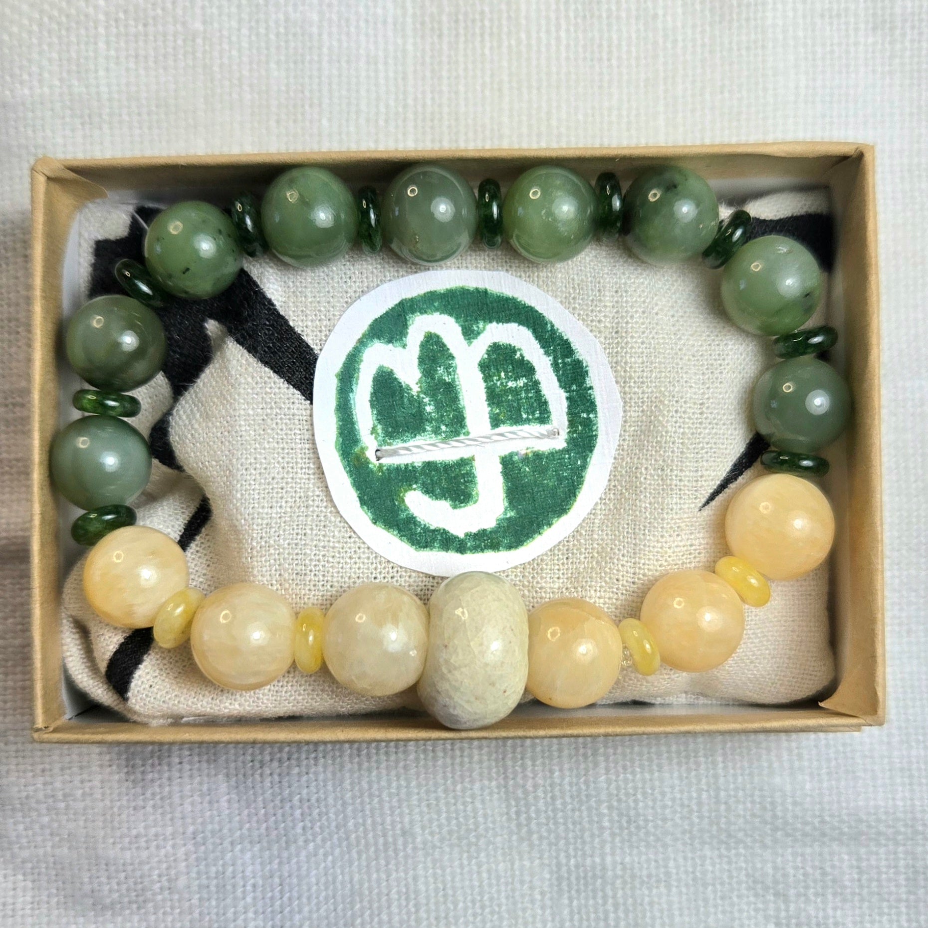 A beaded bracelet displayed inside a light wood gift box on a beige fabric lining. The bracelet features green stone beads on the upper half and pale cream/yellow stone beads on the lower half, separated by small yellow spacer beads.