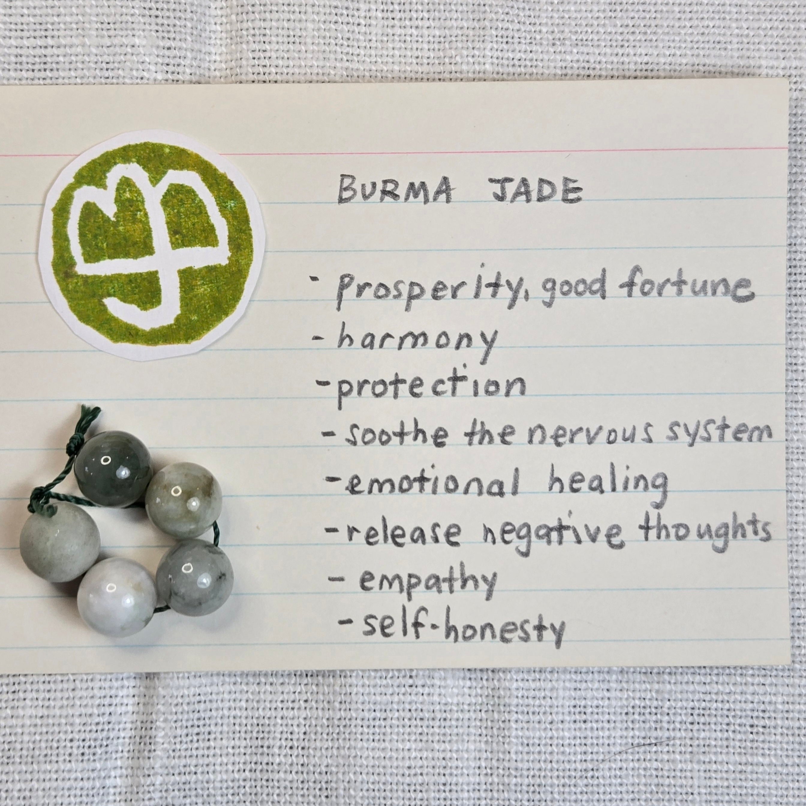 A handmade Burma Jade beaded garland with gray and white marbled beads on a string, displayed next to a handwritten note labeled 'BURMA JADE' with a green gemstone logo, set on a textured white fabric background.
