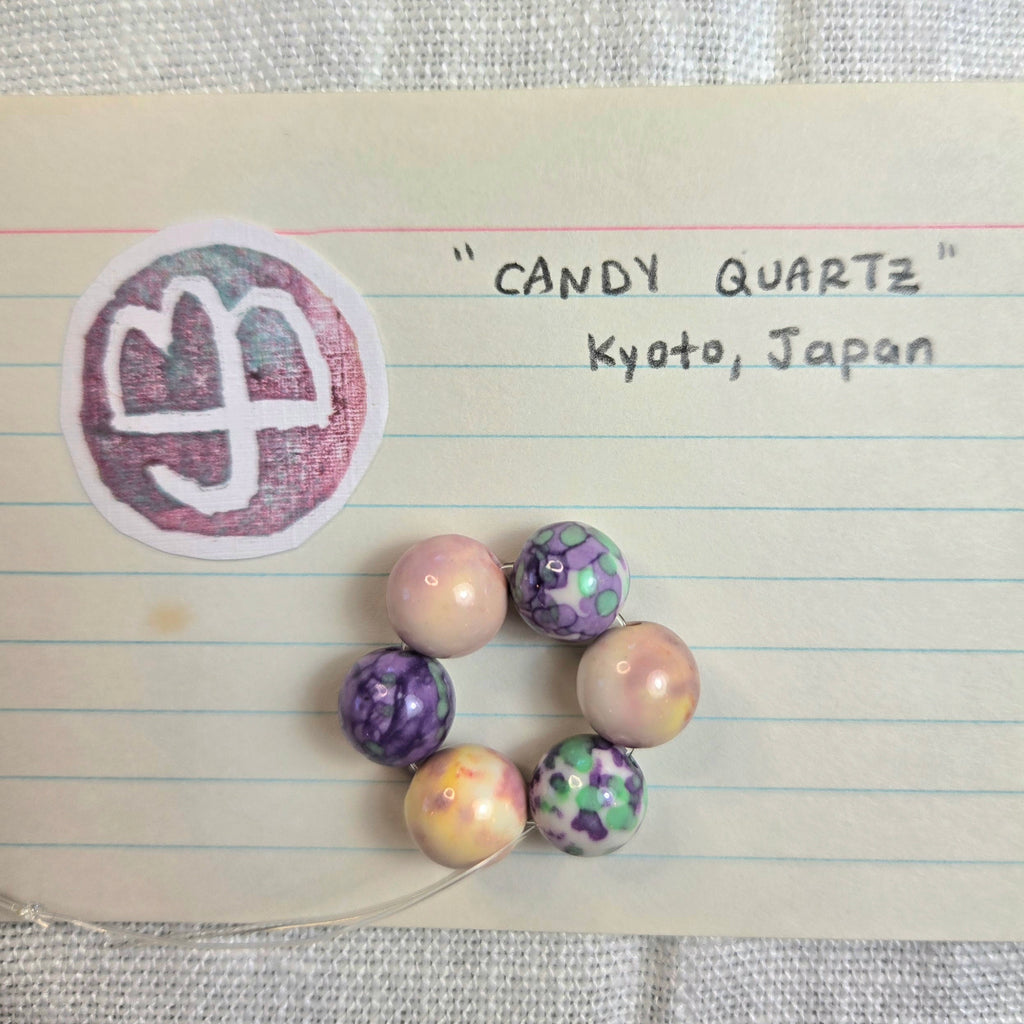 A handmade beaded bracelet made of seven marbled and patterned quartz beads in purple, pink, green, and cream, displayed on a piece of lined notebook paper with handwritten text reading 'CANDY QUARTZ Kyoto, Japan' and a circular stamp.