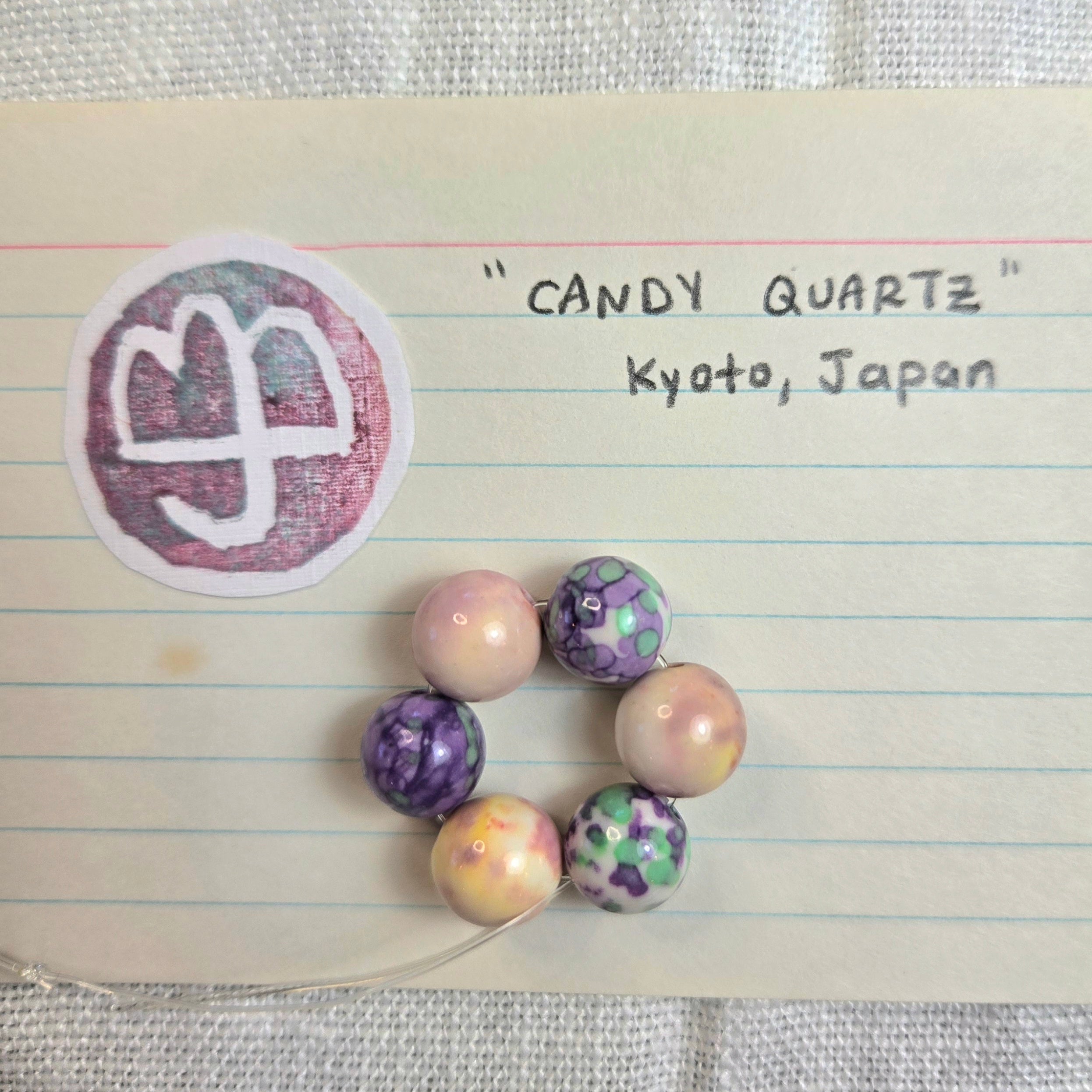 A handmade beaded bracelet made of seven marbled and patterned quartz beads in purple, pink, green, and cream, displayed on a piece of lined notebook paper with handwritten text reading 'CANDY QUARTZ Kyoto, Japan' and a circular stamp.