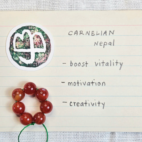 A logo on a green textured background, displayed next to a small ring of orange-red beads on lined paper with handwritten text reading 'CARNELIAN Nepal - boost vitality - motivation - creativity'.