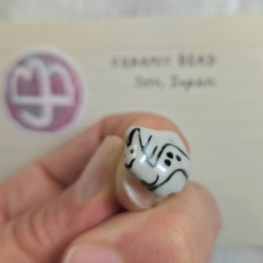 A hand holding a small white ceramic bead with black hand-painted patterns, with a blurred card in the background reading 'CERAMIC BEAD' and 'Jpn, Japan'.