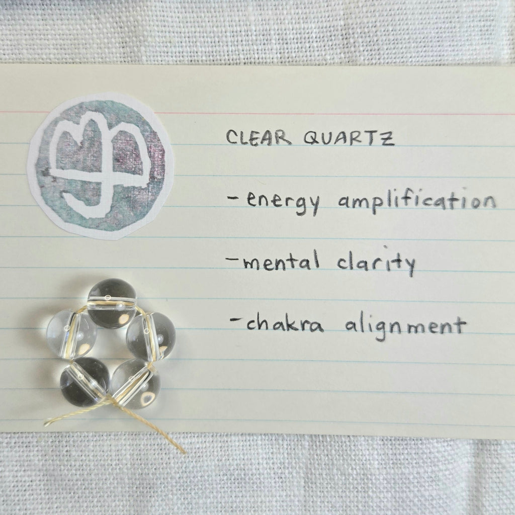 Five clear quartz beads arranged in a chakra-like hexagon on a lined note card next to a circular clear quartz stamp, with handwritten text listing benefits: energy amplification, mental clarity, and chakra alignment.