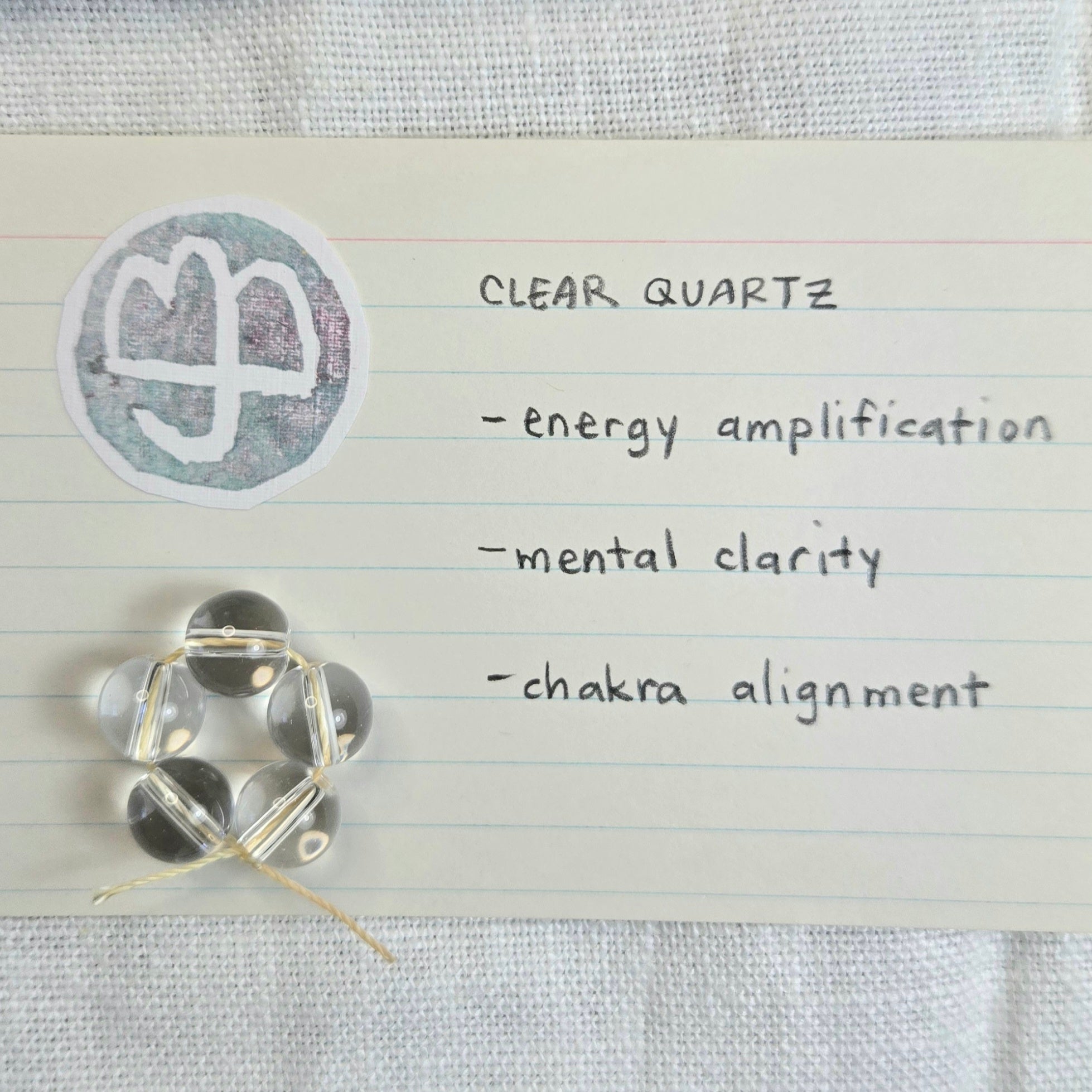 Five clear quartz beads arranged in a chakra-like hexagon on a lined note card next to a circular clear quartz stamp, with handwritten text listing benefits: energy amplification, mental clarity, and chakra alignment.