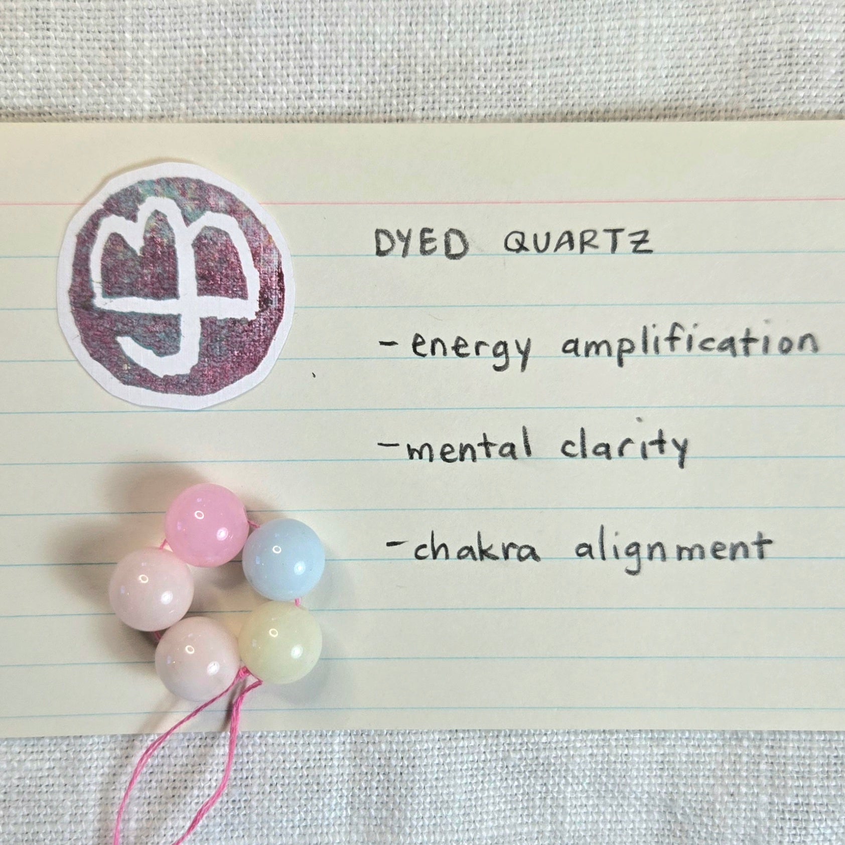 A small set of pastel dyed quartz beads on a pink string, placed on lined paper next to a sticker with a white symbol and handwritten notes reading 'DYED QUARTZ - energy amplification - mental clarity - chakra alignment'.