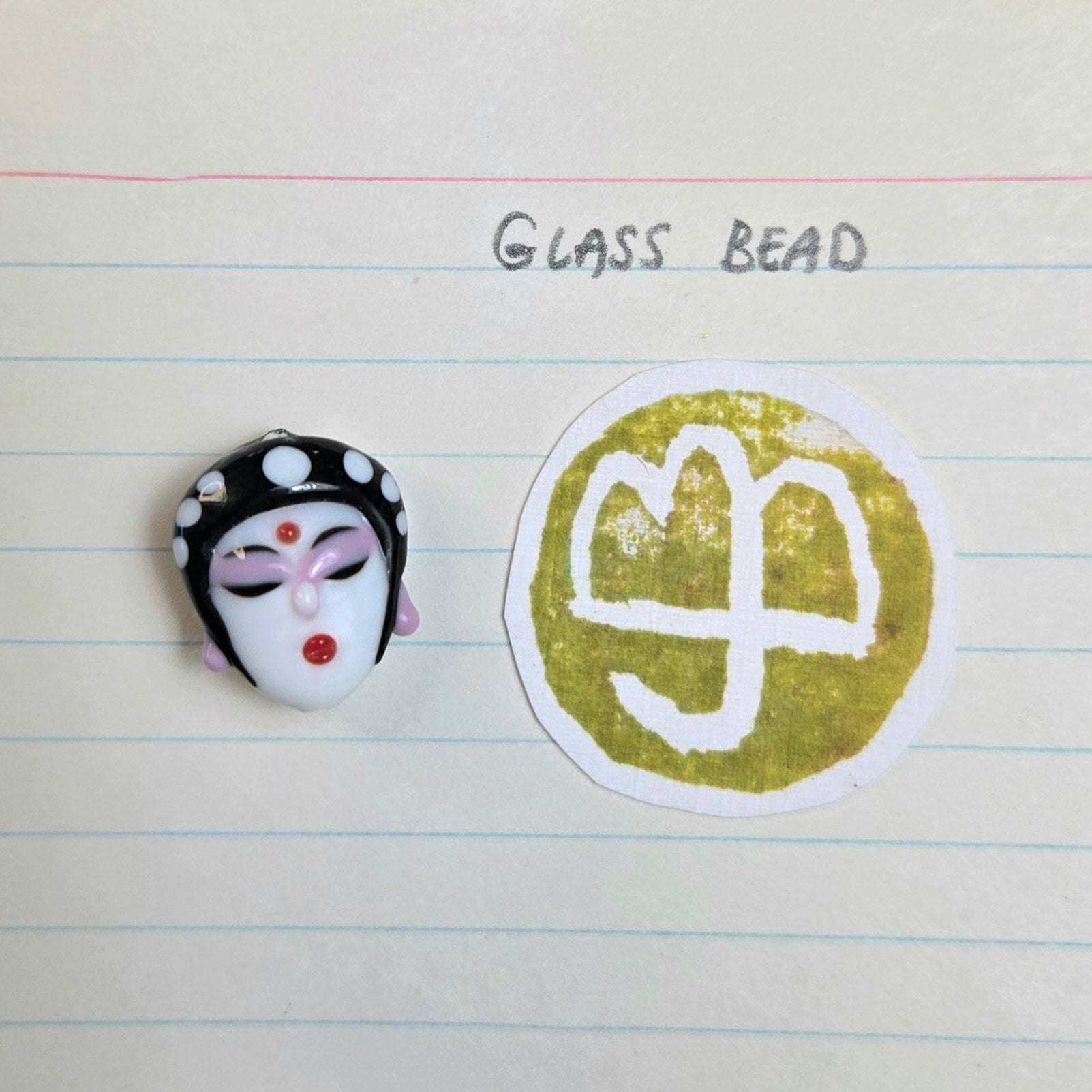 A handmade glass bead shaped like a traditional Chinese opera mask with black, white, and pink details, displayed next to a circular gold foil stamp on a lined notebook page with the handwritten text 'GLASS BEAD'.