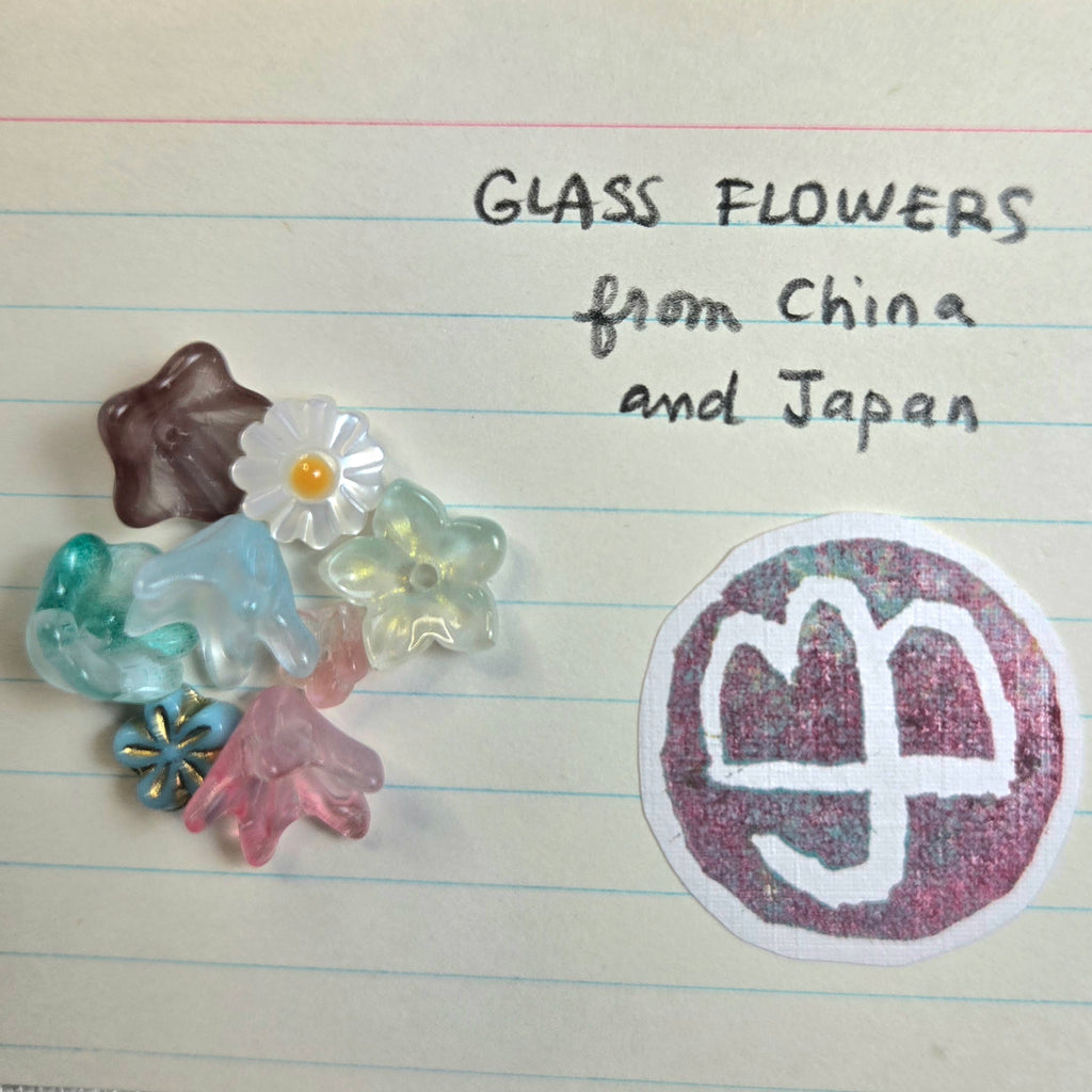 A collection of colorful translucent glass flowers and small decorative glass pieces arranged on lined paper next to handwritten text reading 'GLASS FLOWERS from China and Japan' and a circular logo.