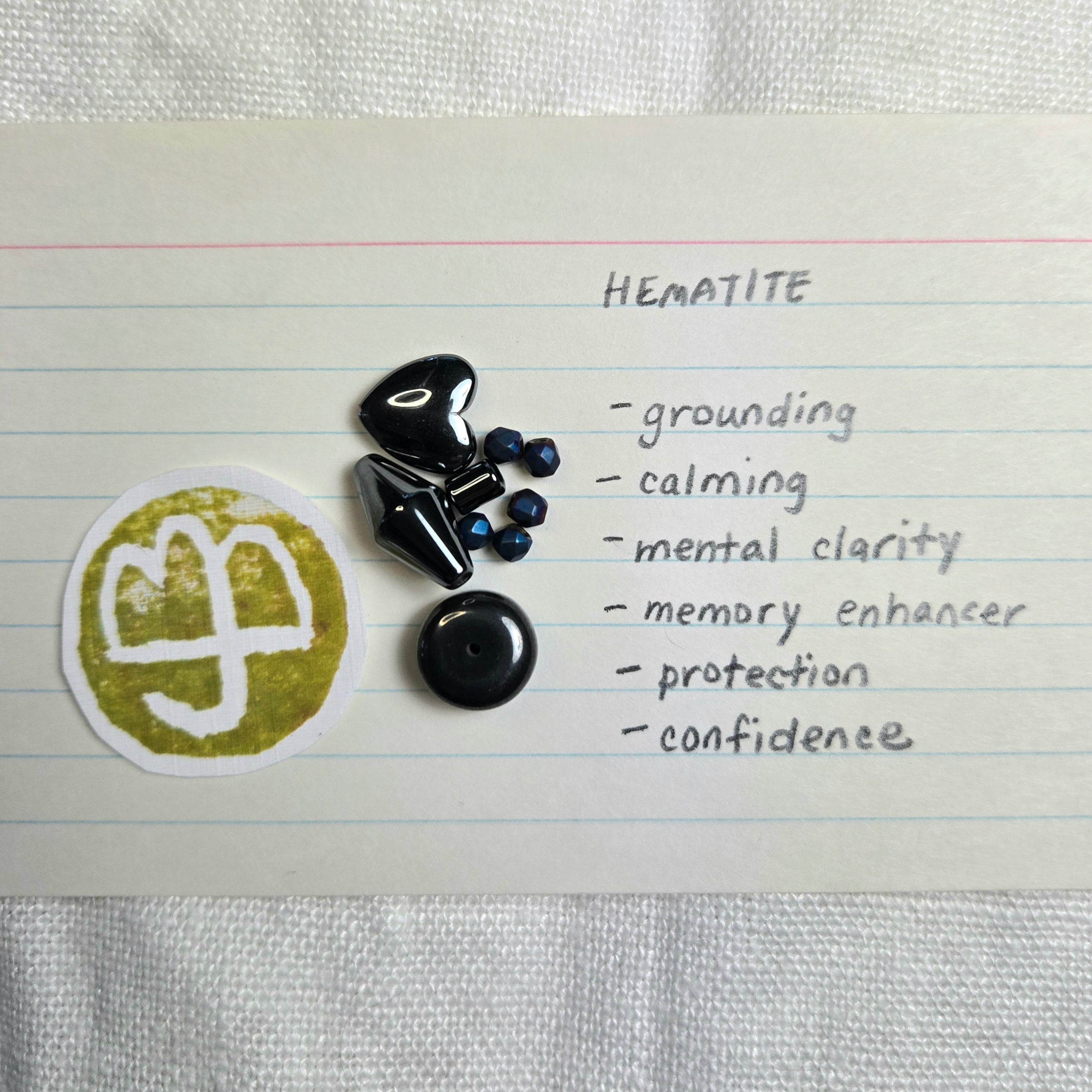 A flat-lay of black hematite crystals and a gold-colored metal charm on lined paper next to a handwritten list titled 'HEMATITE' with positive attributes like grounding, calming, mental clarity, and protection.