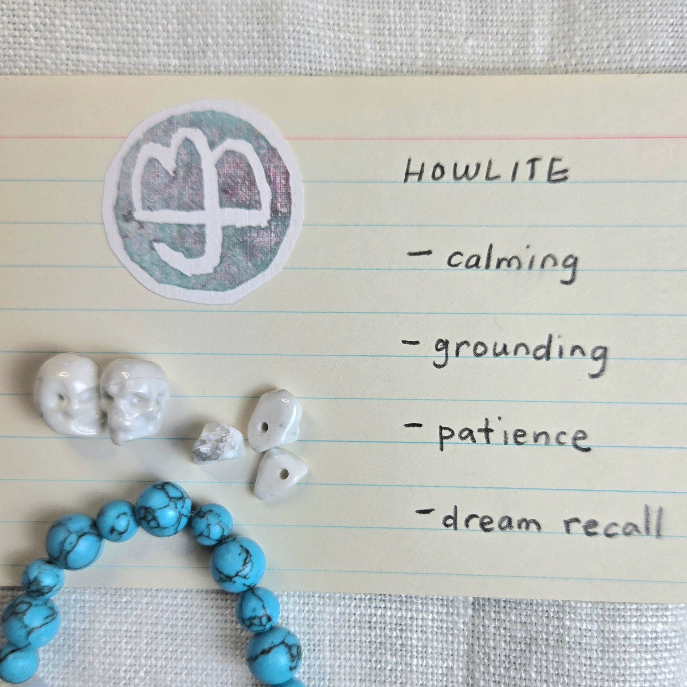 A styled flat-lay of handmade jewelry and crystals on lined paper next to a handwritten list. The items include a blue beaded bracelet, white skull beads, raw turquoise chips, and a circular sticker with a white symbol on a gray-green background.