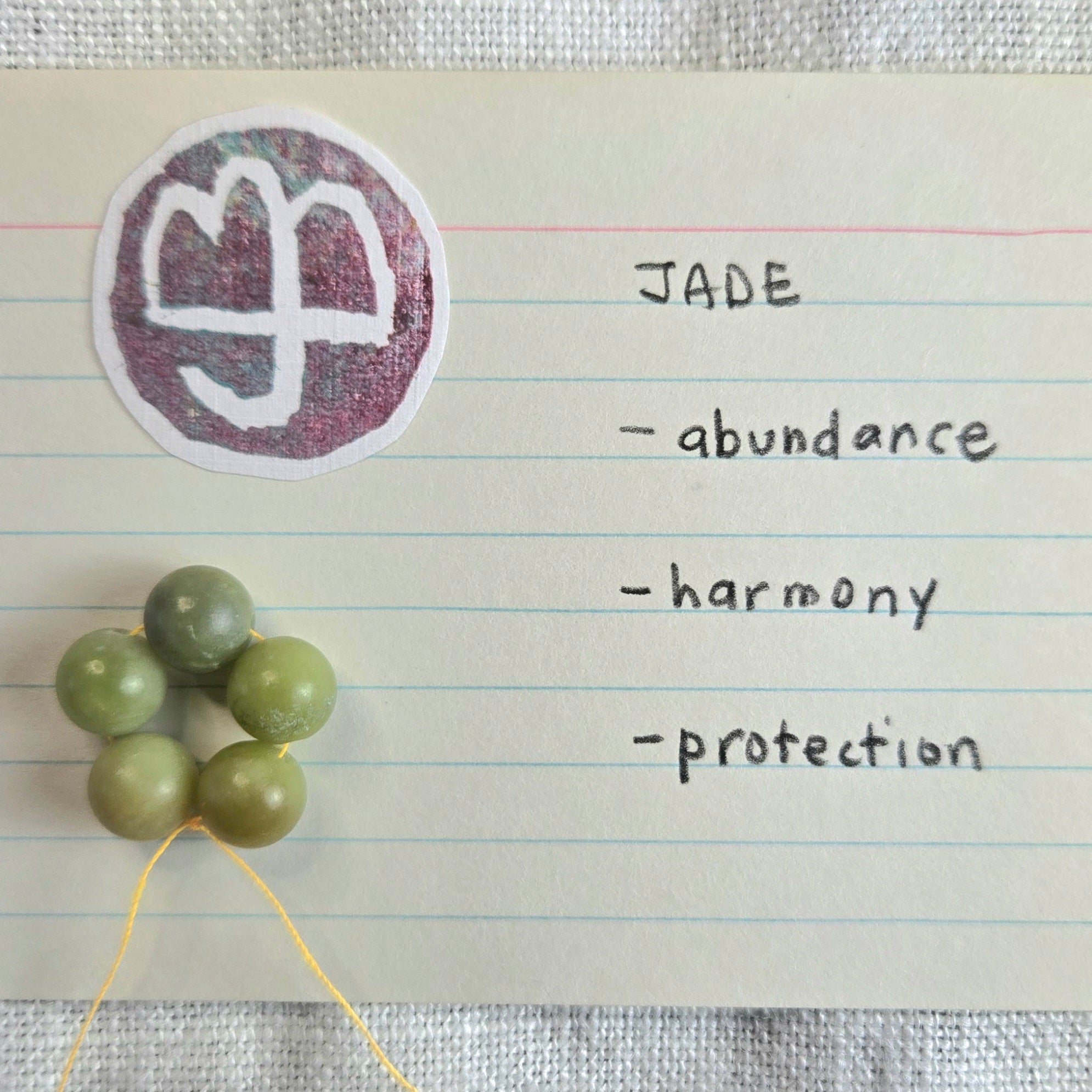 A close-up of handmade green jade beads strung on a yellow thread, placed next to a handwritten note on lined paper. The note features a white symbol inside a purple circle with the word 'JADE' and hand-written qualities: 'abundance', 'harmony', and 'protection'.