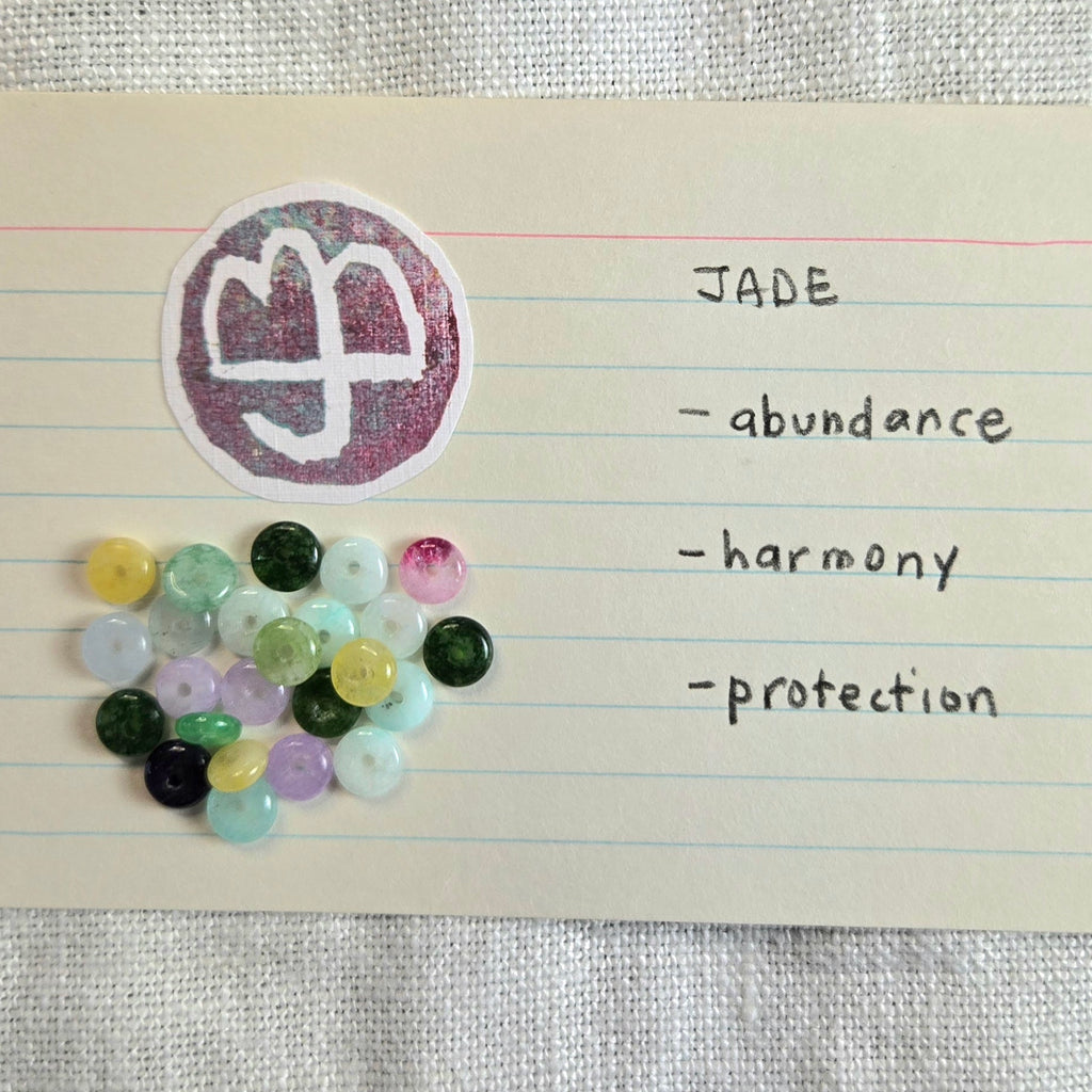 A sheet of lined paper with a handwritten note titled 'JADE' listing qualities 'abundance', 'harmony', and 'protection'. A circular sticker with a white symbol on a pink glitter background is shown above a pile of small round gemstone beads in assorted pastel and dark colors, all placed on a textured fabric surface.