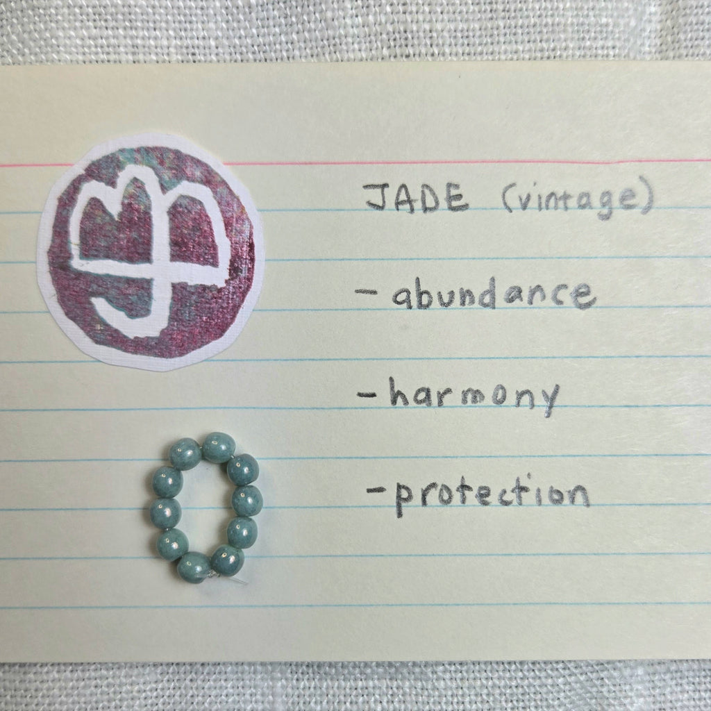 A handmade beaded bracelet made of light blue-green round beads, placed on a piece of lined notebook paper next to a circular sticker with a white symbol and handwritten notes reading 'JADE (vintage)', '-abundance', '-harmony', '-protection'.