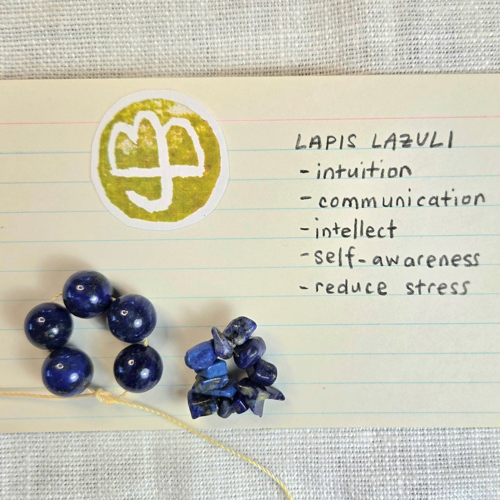 Lapis lazuli beads and a chunky raw stone cluster displayed on lined paper next to a circular stamp with a white semicolon symbol and handwritten notes reading 'LAPIS LAZULI - intuition - communication - intellect - self-awareness - reduce stress'.