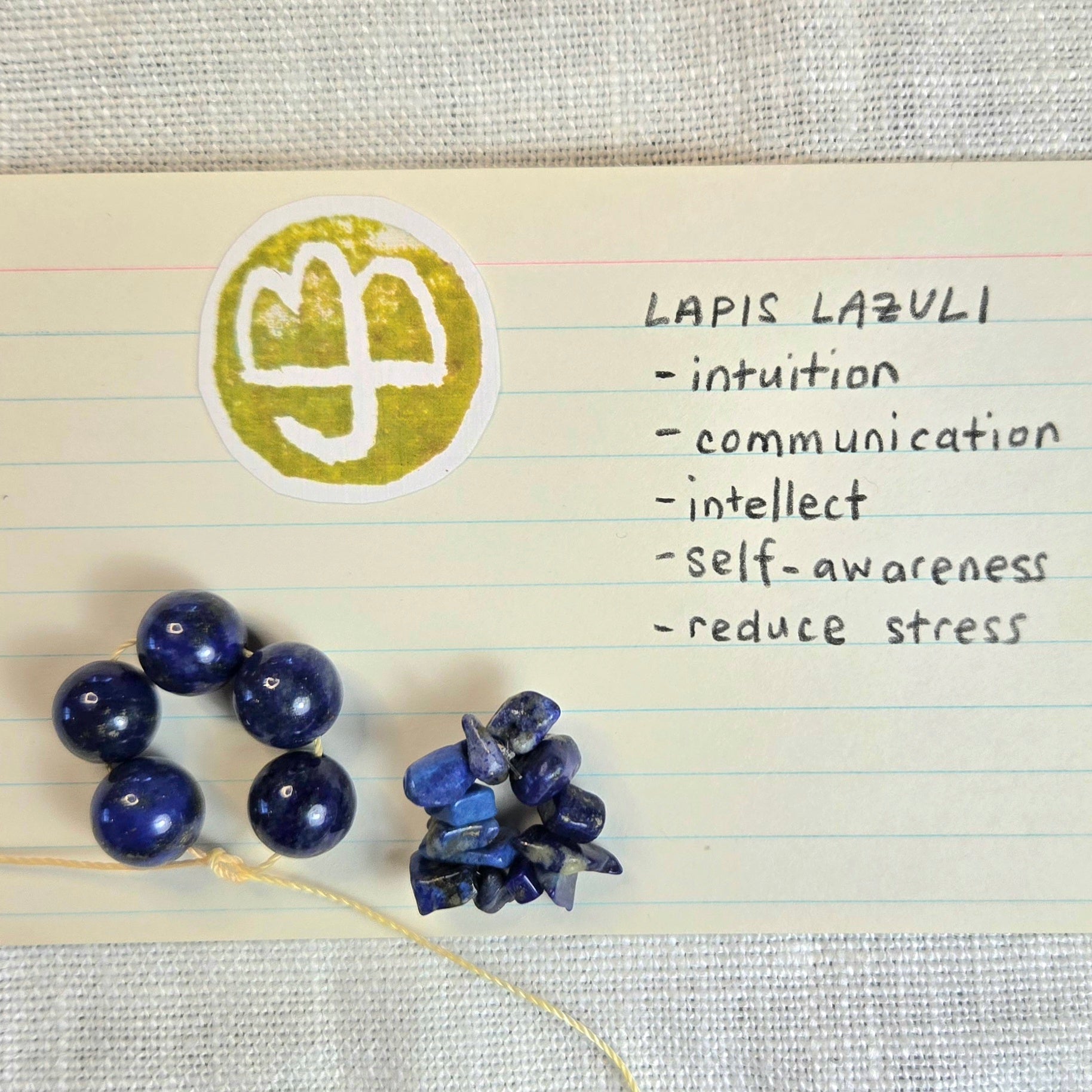 Lapis lazuli beads and a chunky raw stone cluster displayed on lined paper next to a circular stamp with a white semicolon symbol and handwritten notes reading 'LAPIS LAZULI - intuition - communication - intellect - self-awareness - reduce stress'.