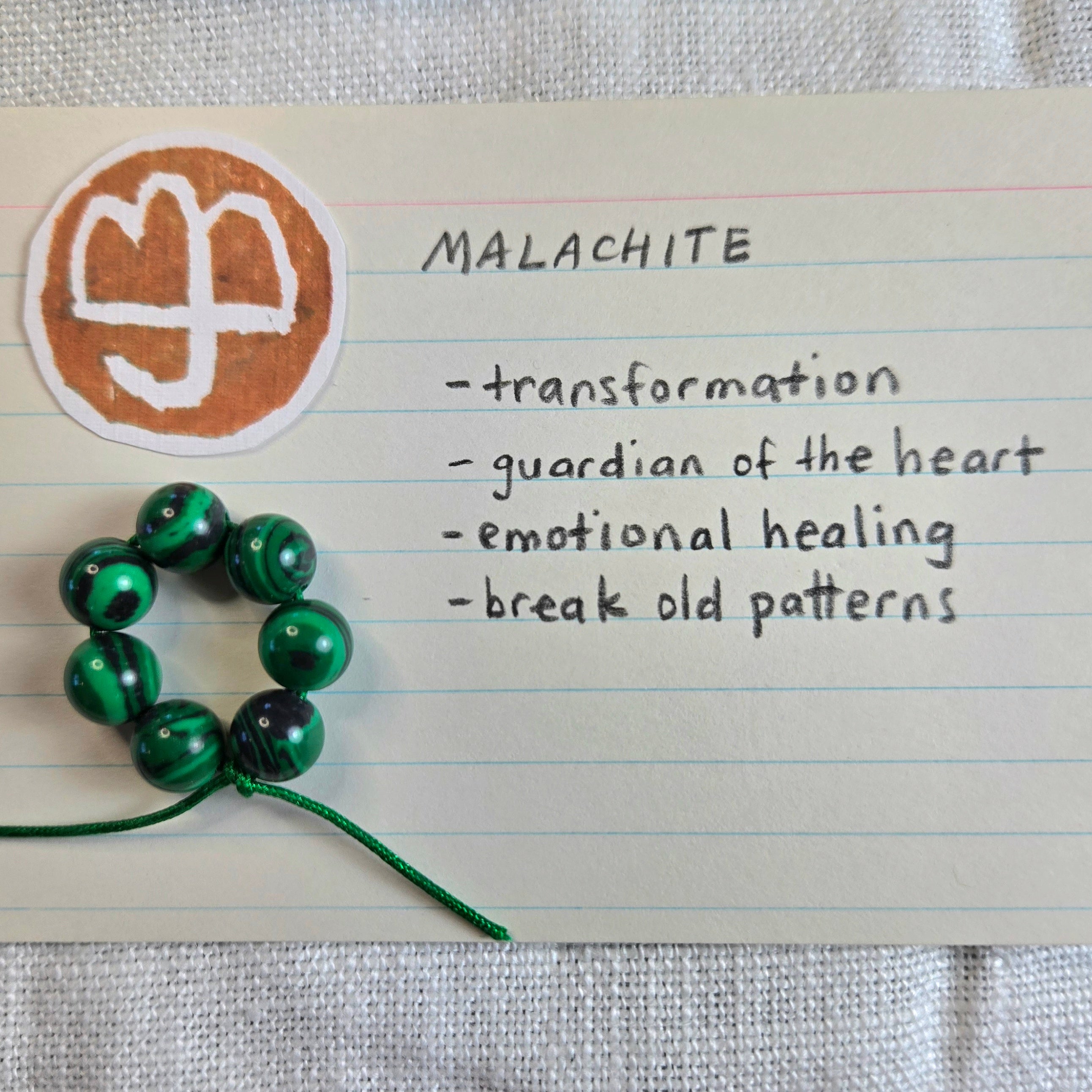 A handmade beaded bracelet made of green and black malachite beads on a string, placed next to a handwritten note card labeled 'MALACHITE' with a chakra symbol and descriptive text.