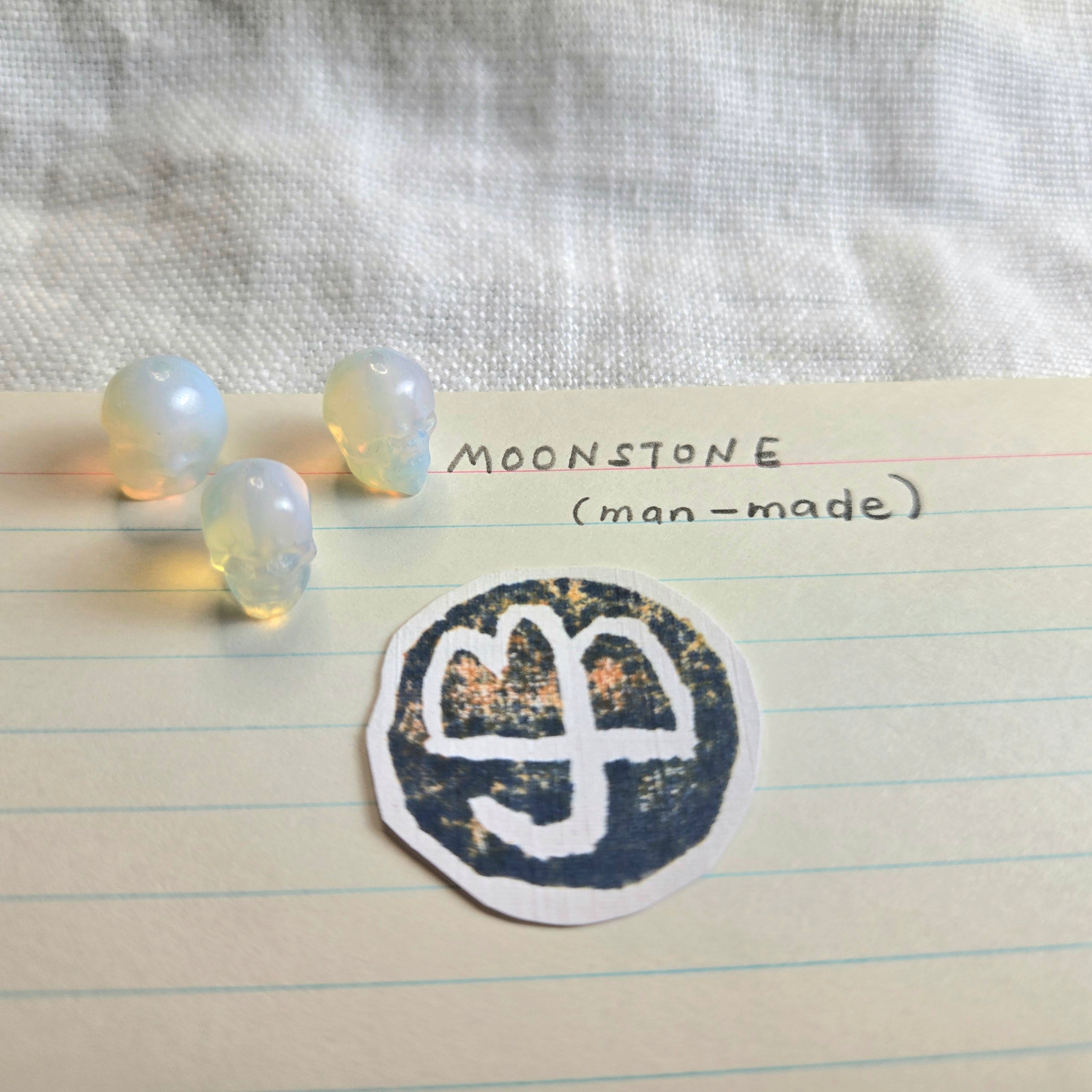 Three small iridescent moonstone beads and a circular stamped stone seal on lined paper with handwritten text reading 'MOONSTONE (man-made)'.
