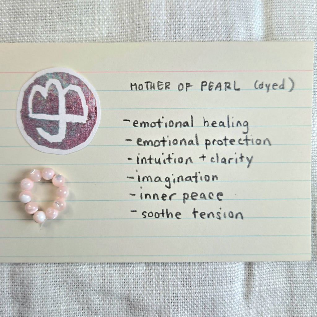 A small pink beaded ring displayed on a cream lined note card next to a circular sticker with a white symbol on a dusty purple background. Handwritten text on the card reads 'MOTHER OF PEARL (dyed)' and lists qualities like emotional healing, intuition, and inner peace.