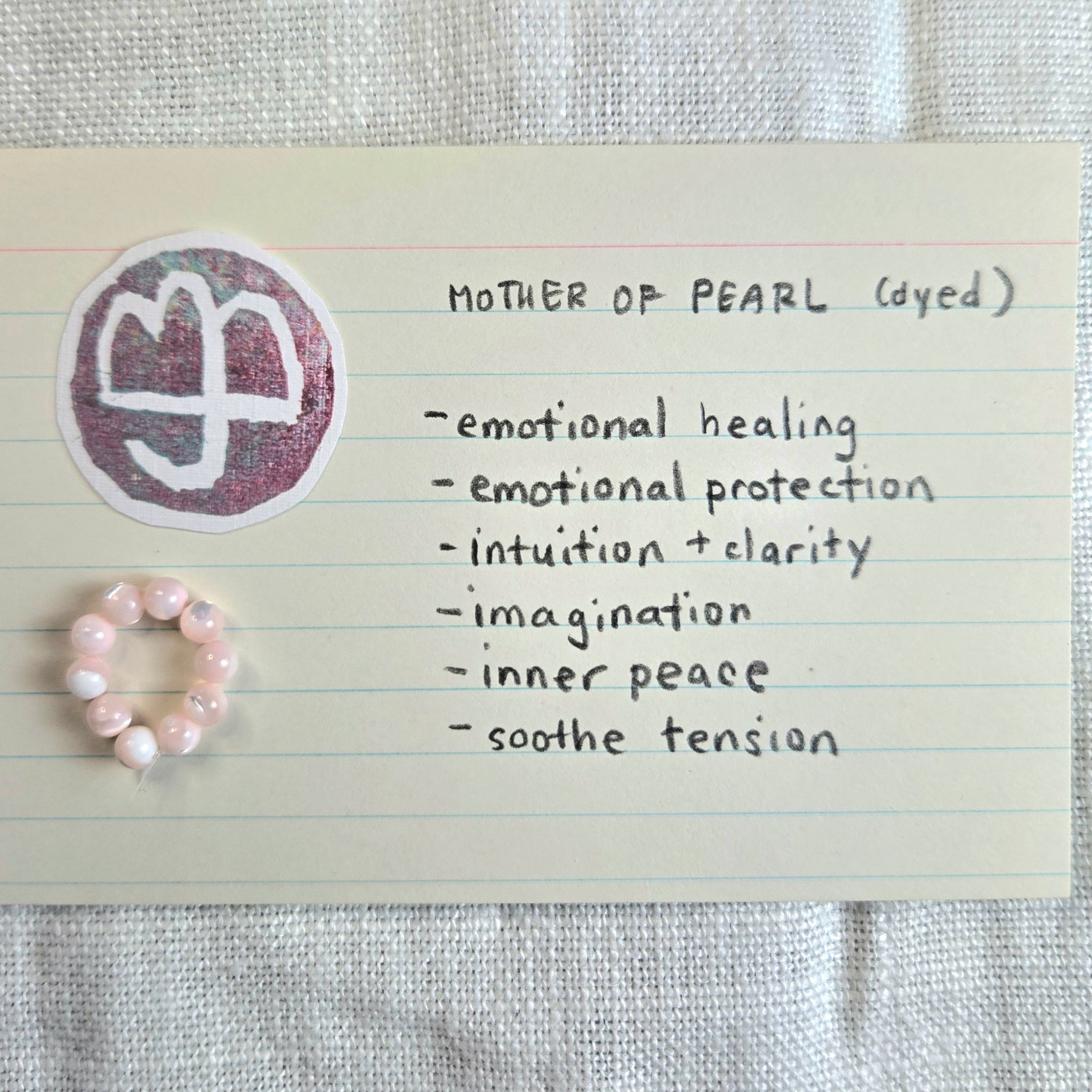 A small pink beaded ring displayed on a cream lined note card next to a circular sticker with a white symbol on a dusty purple background. Handwritten text on the card reads 'MOTHER OF PEARL (dyed)' and lists qualities like emotional healing, intuition, and inner peace.
