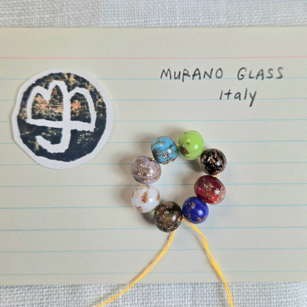 A set of colorful Murano glass beads on a yellow string, displayed on lined paper next to a circular sticker with the word 'MURANO GLASS Italy'.