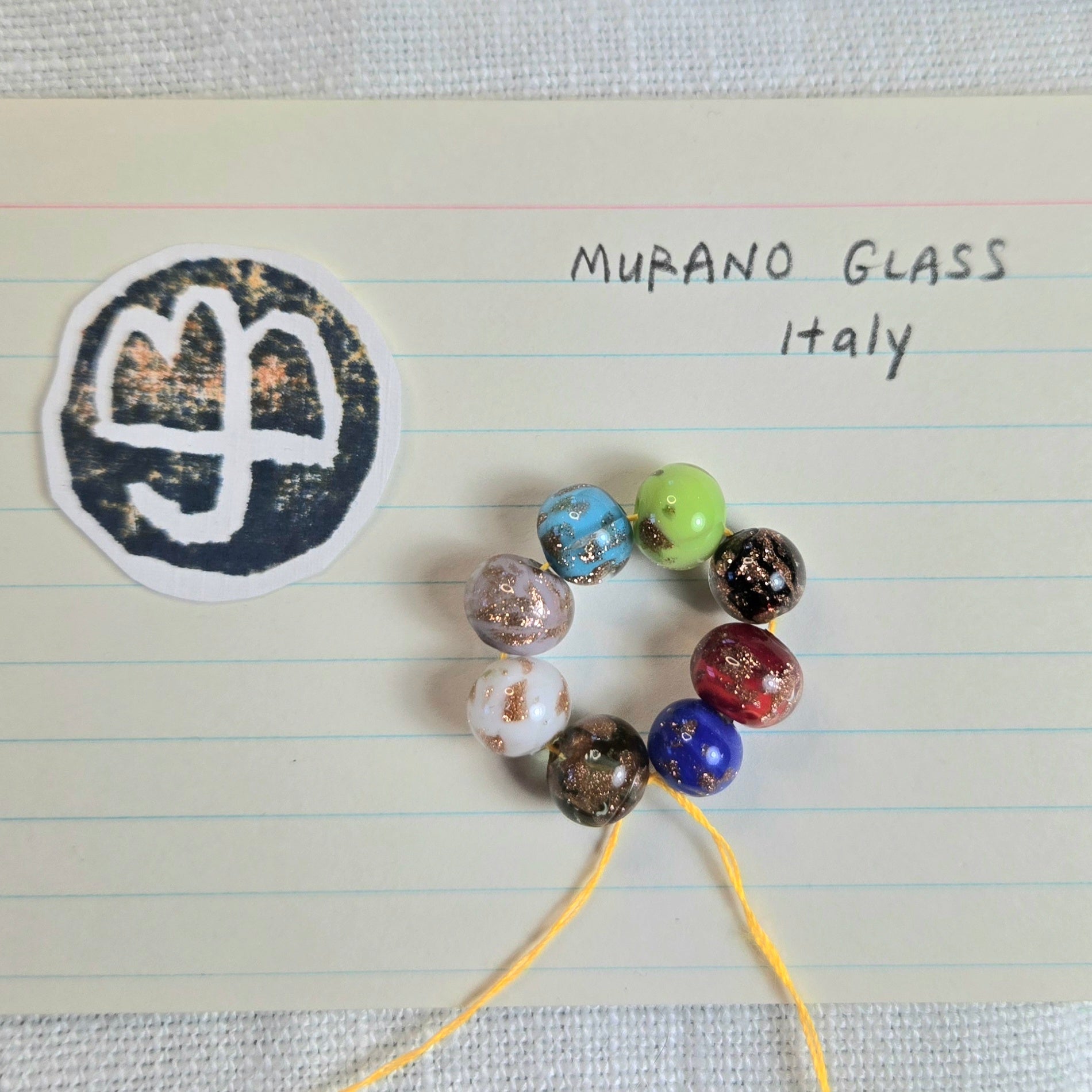 A set of colorful Murano glass beads on a yellow string, displayed on lined paper next to a circular sticker with the word 'MURANO GLASS Italy'.