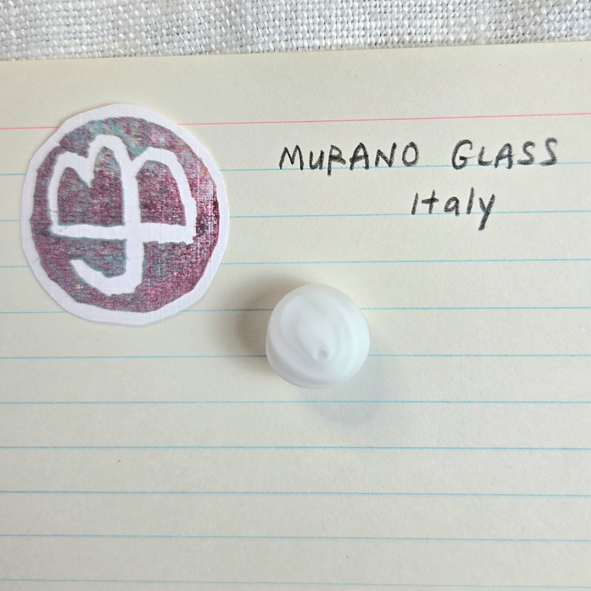 A small white oval glass bead with a textured, frosted finish is displayed on a sheet of lined paper next to a circular sticker featuring the Murano glass logo and the handwritten text 'MURANO GLASS Italy'.