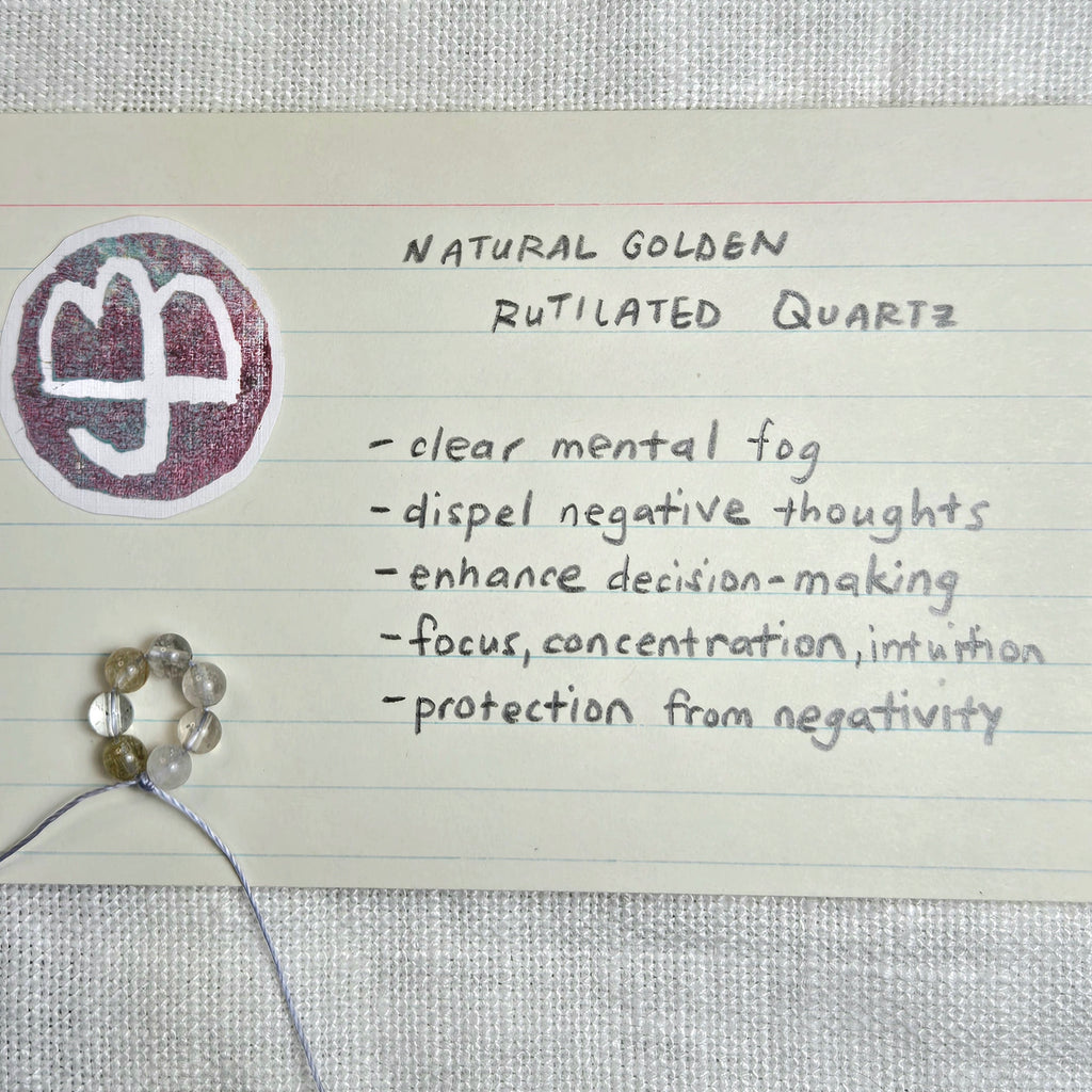 A handwritten note on lined paper describing 'Natural Golden Rutilated Quartz' with benefits listed, alongside a round sticker showing a white symbol on a purple background and several small translucent beads on a string.