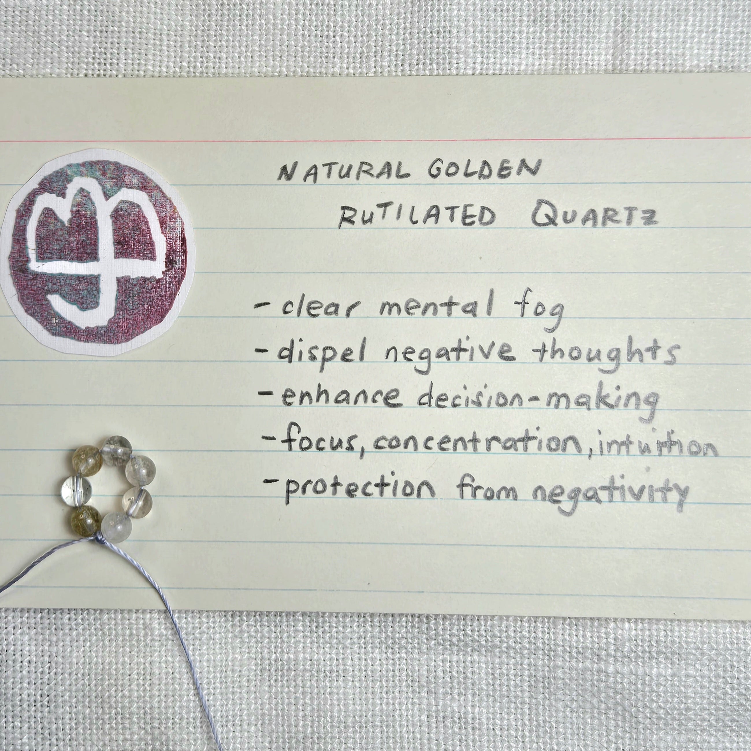 A handwritten note on lined paper describing 'Natural Golden Rutilated Quartz' with benefits listed, alongside a round sticker showing a white symbol on a purple background and several small translucent beads on a string.