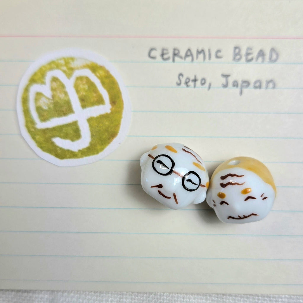 Two small ceramic beads with cartoon faces on a lined paper background next to a green and white logo.