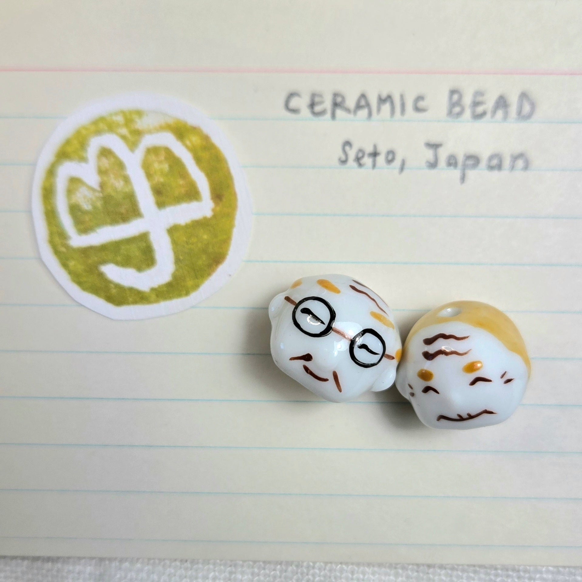 Two small ceramic beads with cartoon faces on a lined paper background next to a green and white logo.