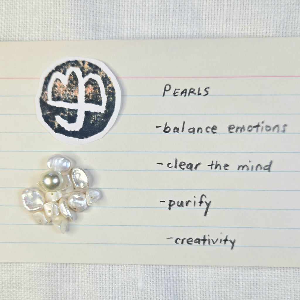 A small cluster of white pearls with one larger pale pearl, placed on a lined index card next to a black circular symbol and handwritten words including 'PEARLS', balance emotions, clear the mind, purify, and creativity