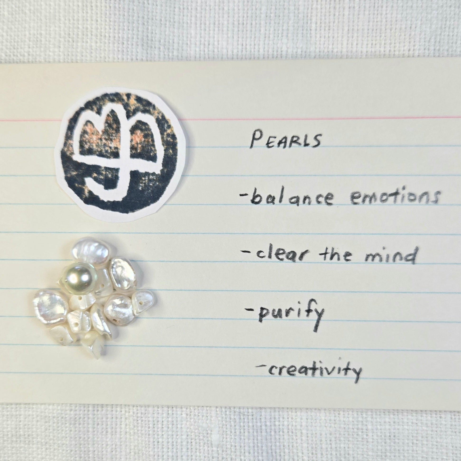 A small cluster of white pearls with one larger pale pearl, placed on a lined index card next to a black circular symbol and handwritten words including 'PEARLS', balance emotions, clear the mind, purify, and creativity