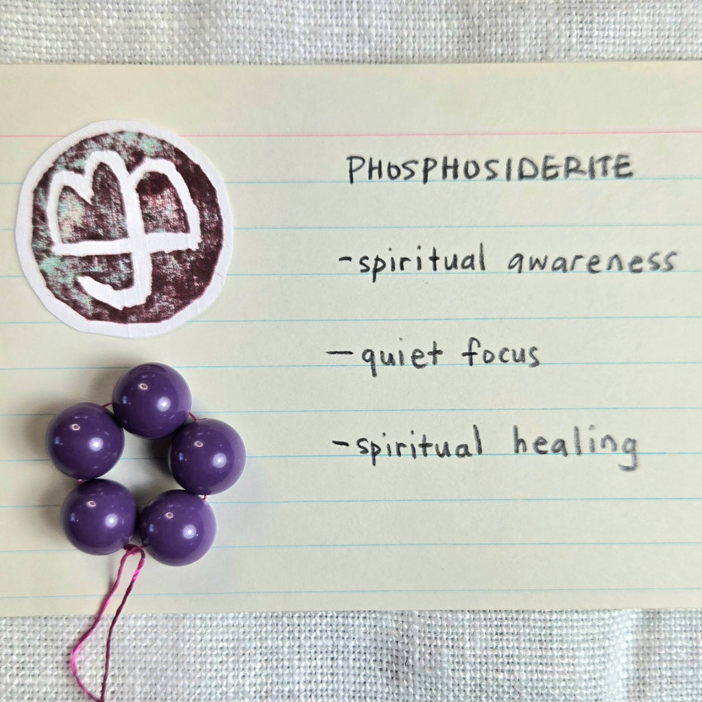 Purple spherical beads on a string arranged on a lined card next to a circular white and brown logo, with handwritten text reading 'PHOSPHOSIDERITE' and descriptors including 'spiritual awareness', 'quiet focus', and 'spiritual healing'.