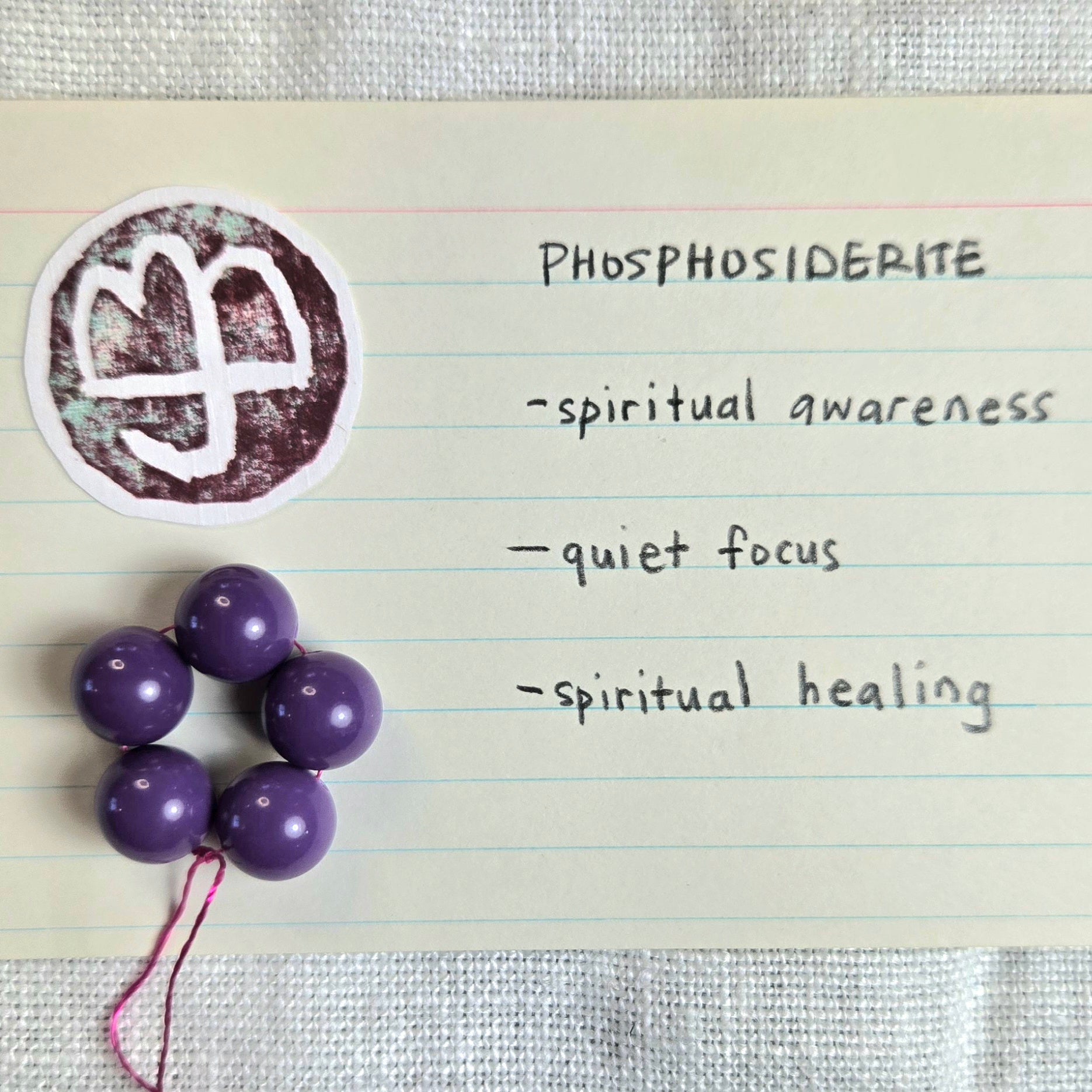 Purple spherical beads on a string arranged on a lined card next to a circular white and brown logo, with handwritten text reading 'PHOSPHOSIDERITE' and descriptors including 'spiritual awareness', 'quiet focus', and 'spiritual healing'.