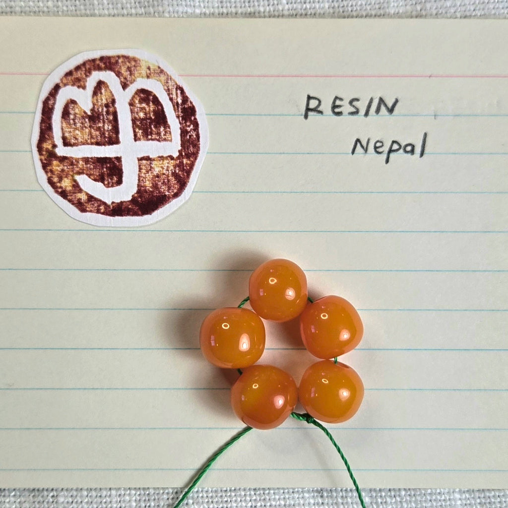 Five glossy amber-orange resin beads strung on a green cord, displayed on lined paper next to a circular stamp of a logo and handwritten text reading 'RESIN Nepal'.