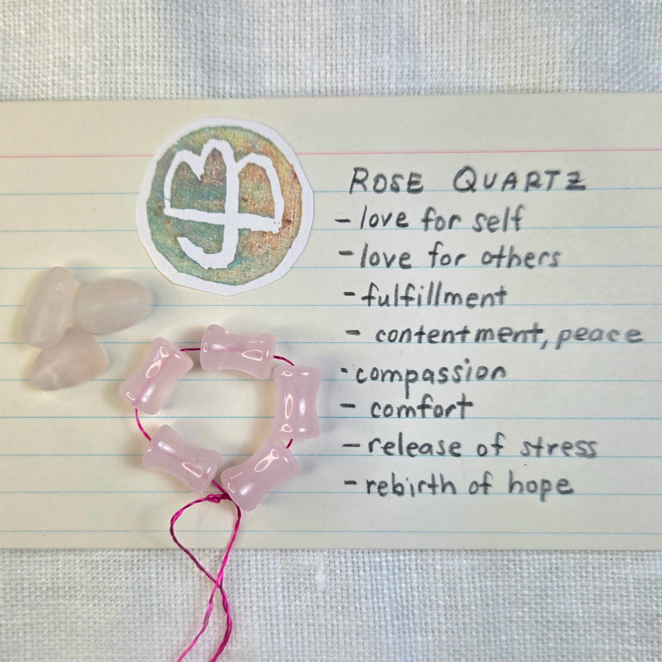 A heart-shaped rosary bead necklace made of pale pink rose quartz beads on a pink cord, displayed on a lined paper card with a handwritten list of rose quartz meanings and a small white crystal cluster.