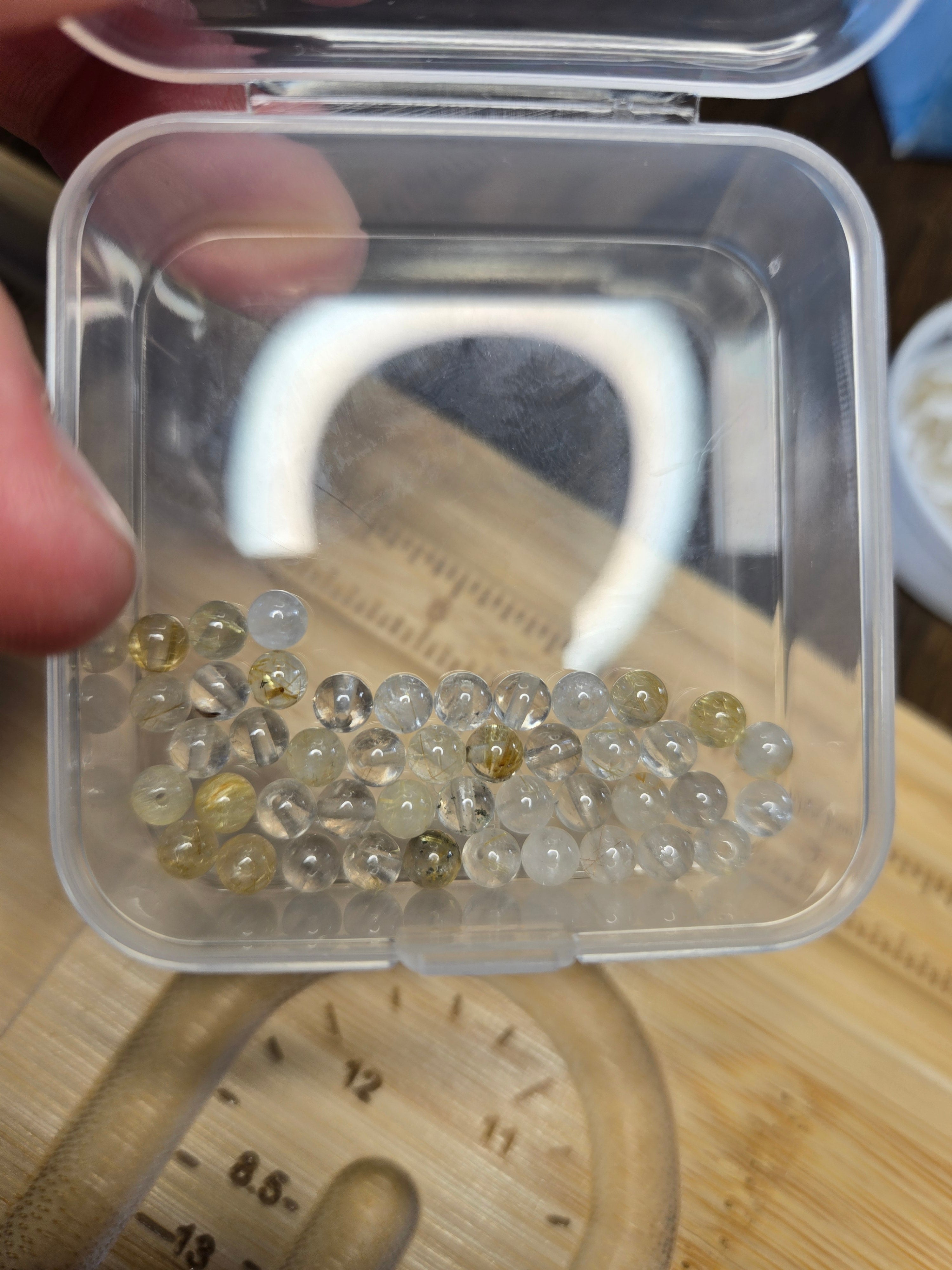 A hand holding a small clear plastic case containing loose round transparent beads with some pale yellow and clear stones, photographed over a wooden surface.