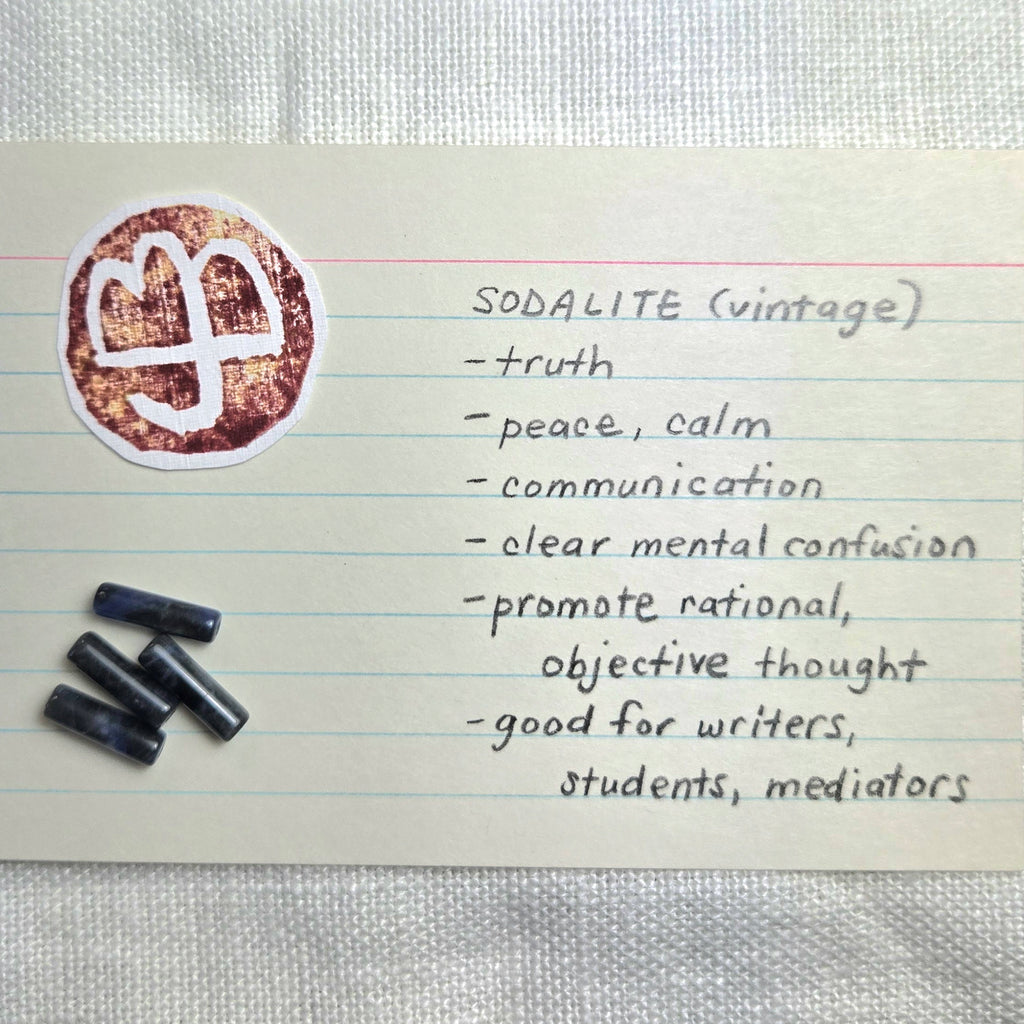 A handwritten note on lined paper describing 'SODALITE (vintage)' with a list of qualities and a round logo sticker, plus three small dark rectangular items placed beside it on a light fabric background.