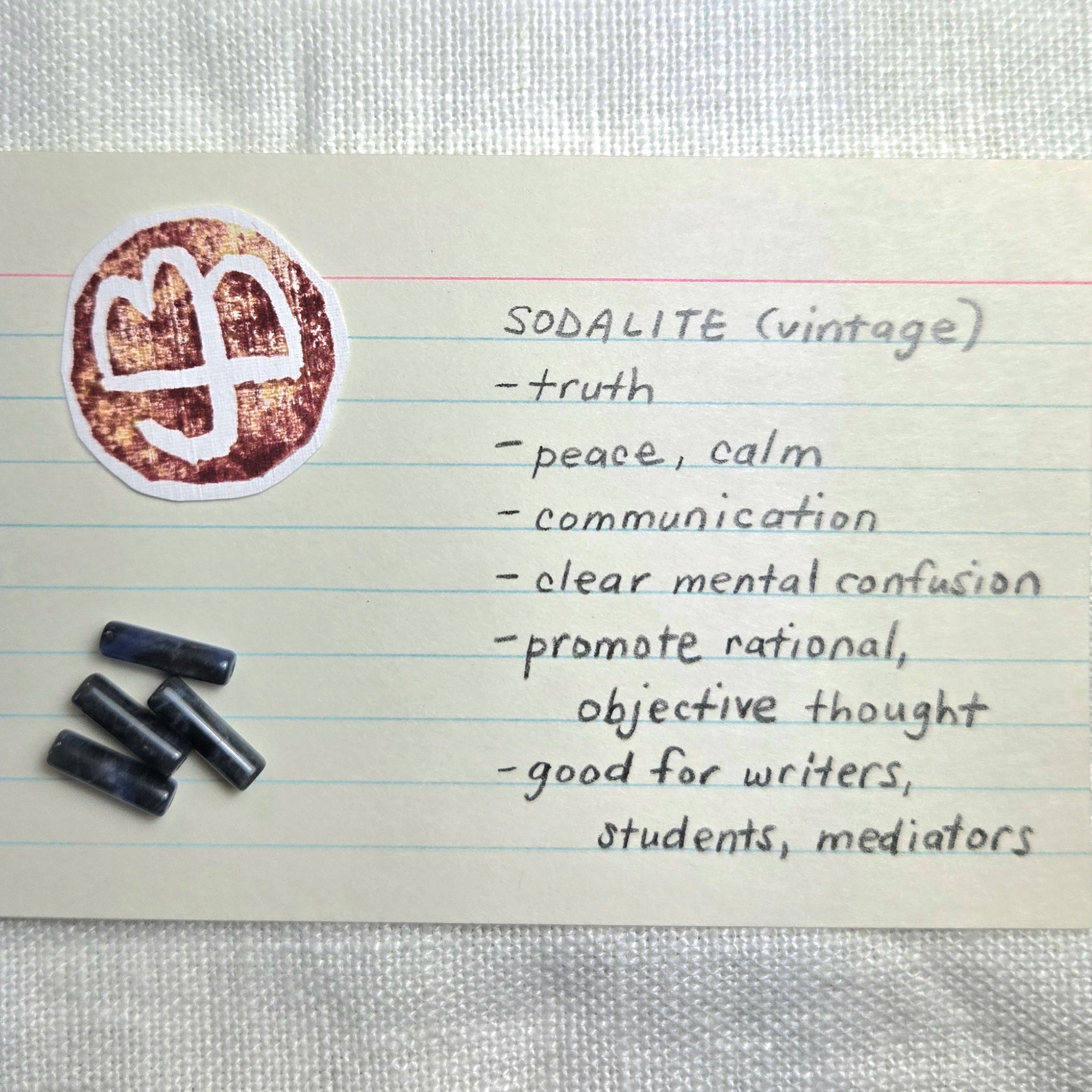 A handwritten note on lined paper describing 'SODALITE (vintage)' with a list of qualities and a round logo sticker, plus three small dark rectangular items placed beside it on a light fabric background.