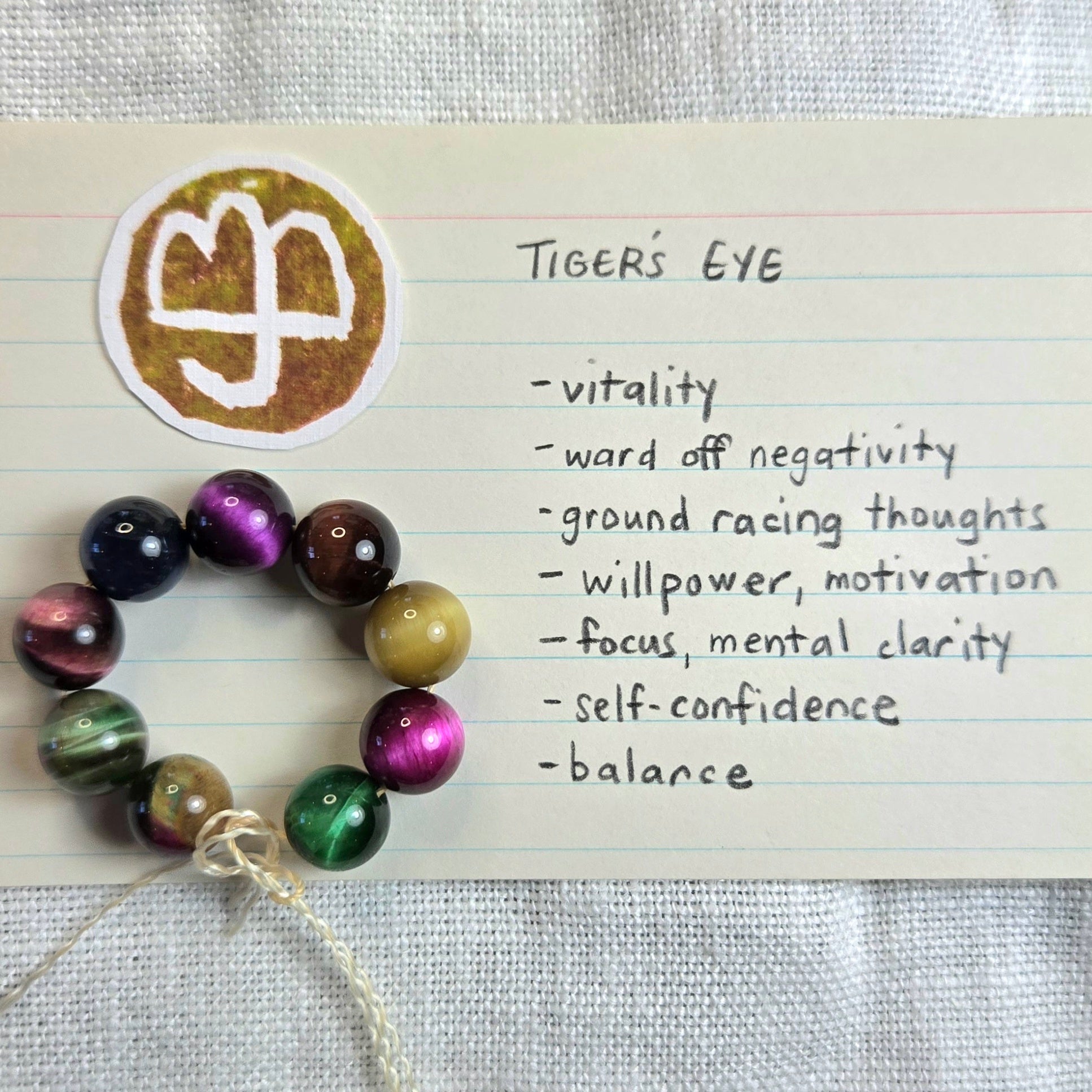 A handmade beaded necklace with tiger's eye beads arranged in a circle on a lined notebook page next to a gold-toned tiger's eye charm and a handwritten list titled 'TIGER'S EYE'.