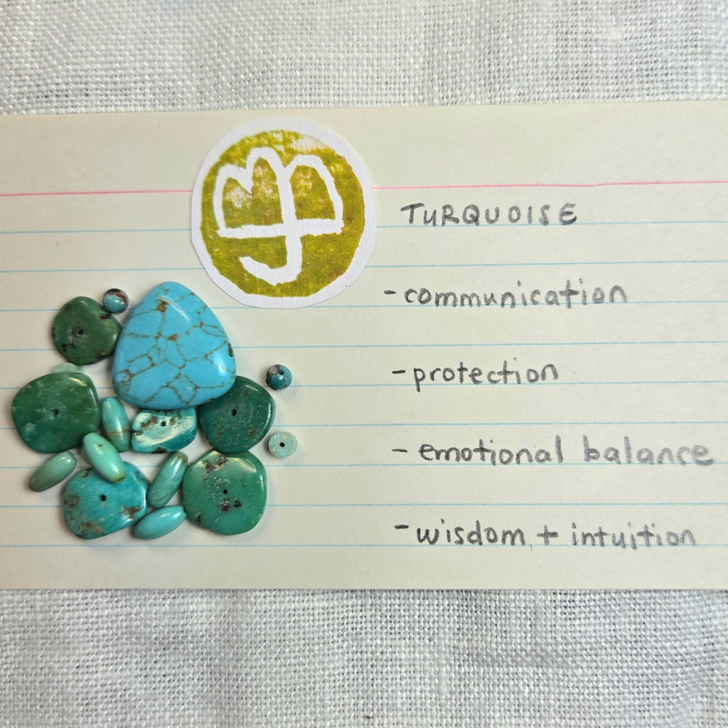 Turquoise gemstone beads and a polished turquoise stone arranged on a lined paper card next to a yellow circular sticker with a white symbol. The card lists meanings for turquoise: communication, protection, emotional balance, and wisdom + intuition.