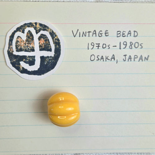 A small glossy yellow vintage bead is displayed on lined paper next to a handwritten note and a circular black-and-teal-and-orange logo. It reads: Vintage bead 1970s to 1980s, Osaka, Japan