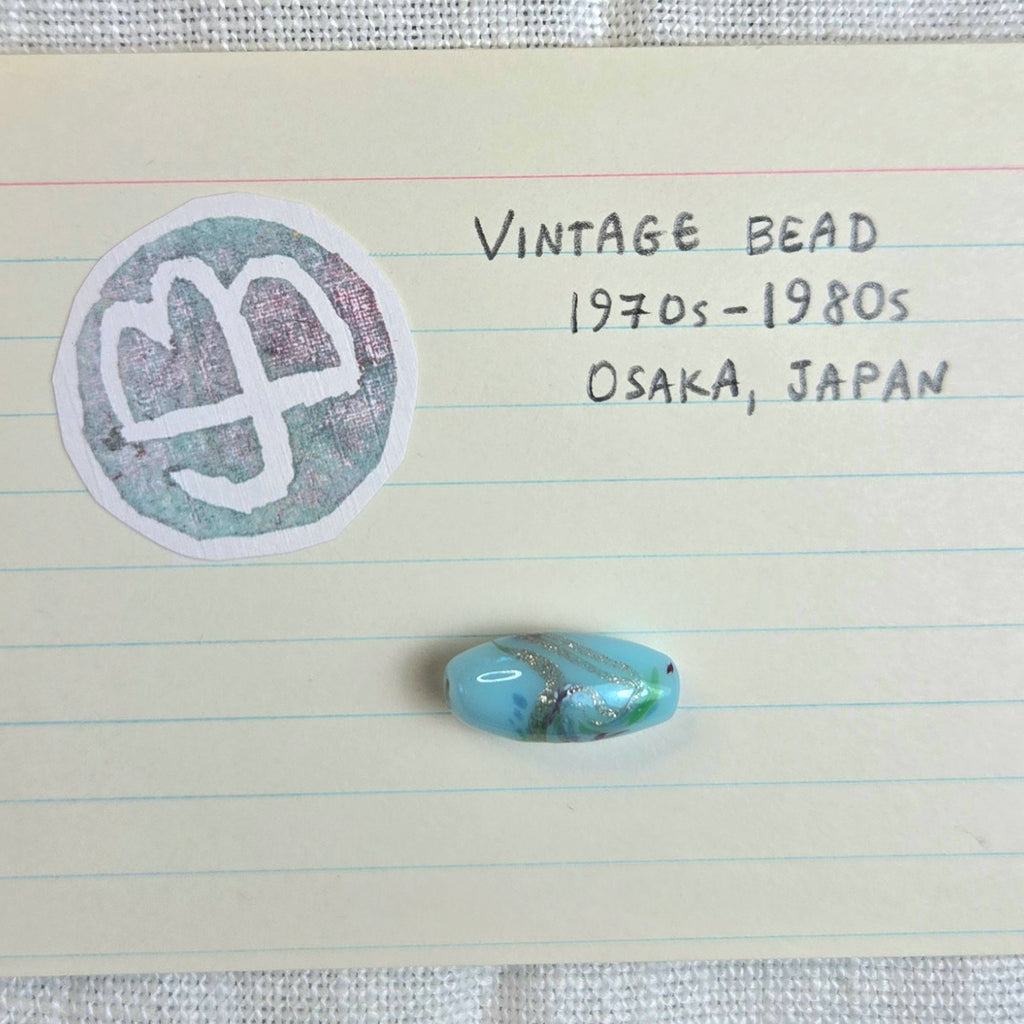 A single oval vintage bead with a light blue and white marbled pattern is displayed on lined paper next to handwritten notes reading 'VINTAGE BEAD 1970s-1980s OSAKA, JAPAN' and a circular stamp.