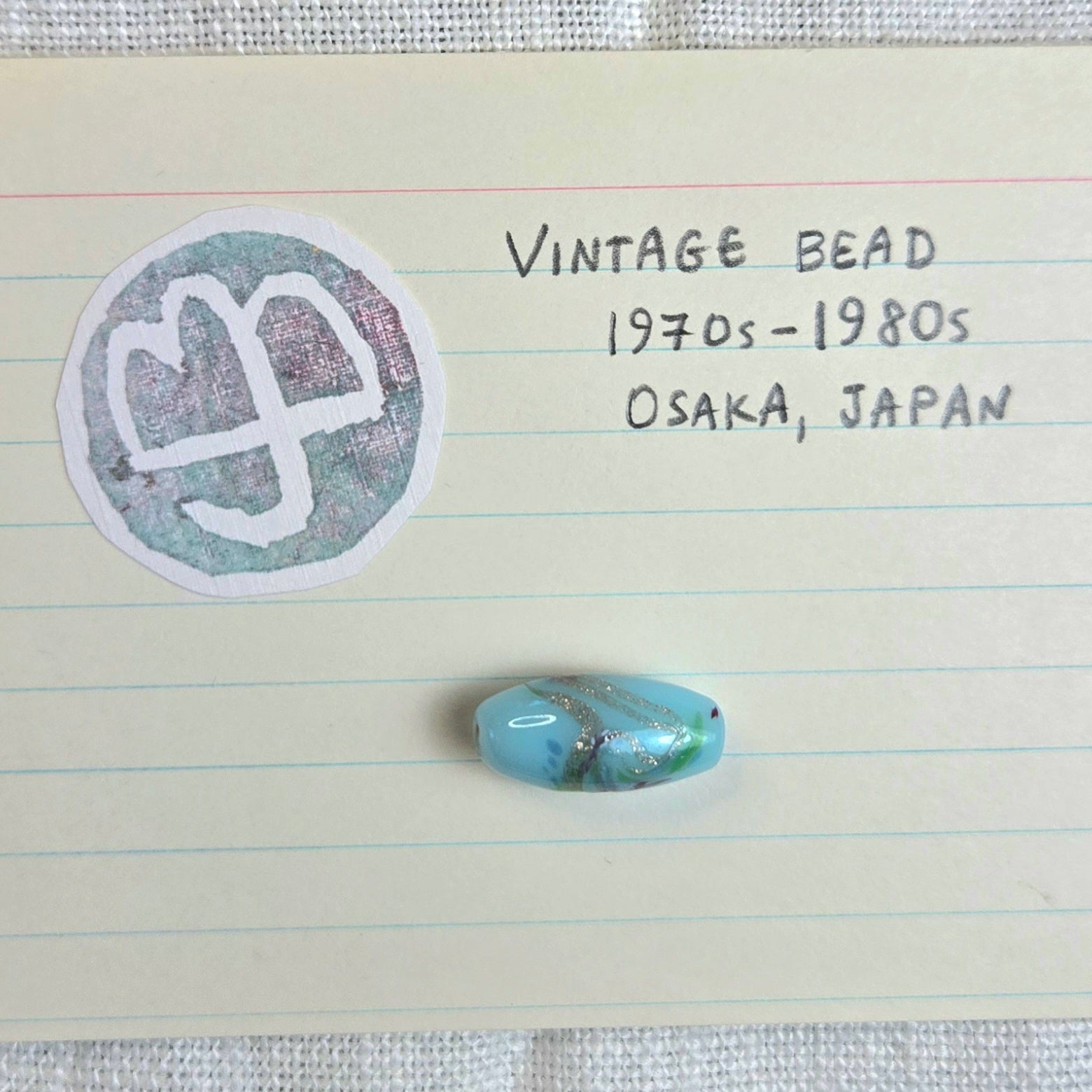 A single oval vintage bead with a light blue and white marbled pattern is displayed on lined paper next to handwritten notes reading 'VINTAGE BEAD 1970s-1980s OSAKA, JAPAN' and a circular stamp.