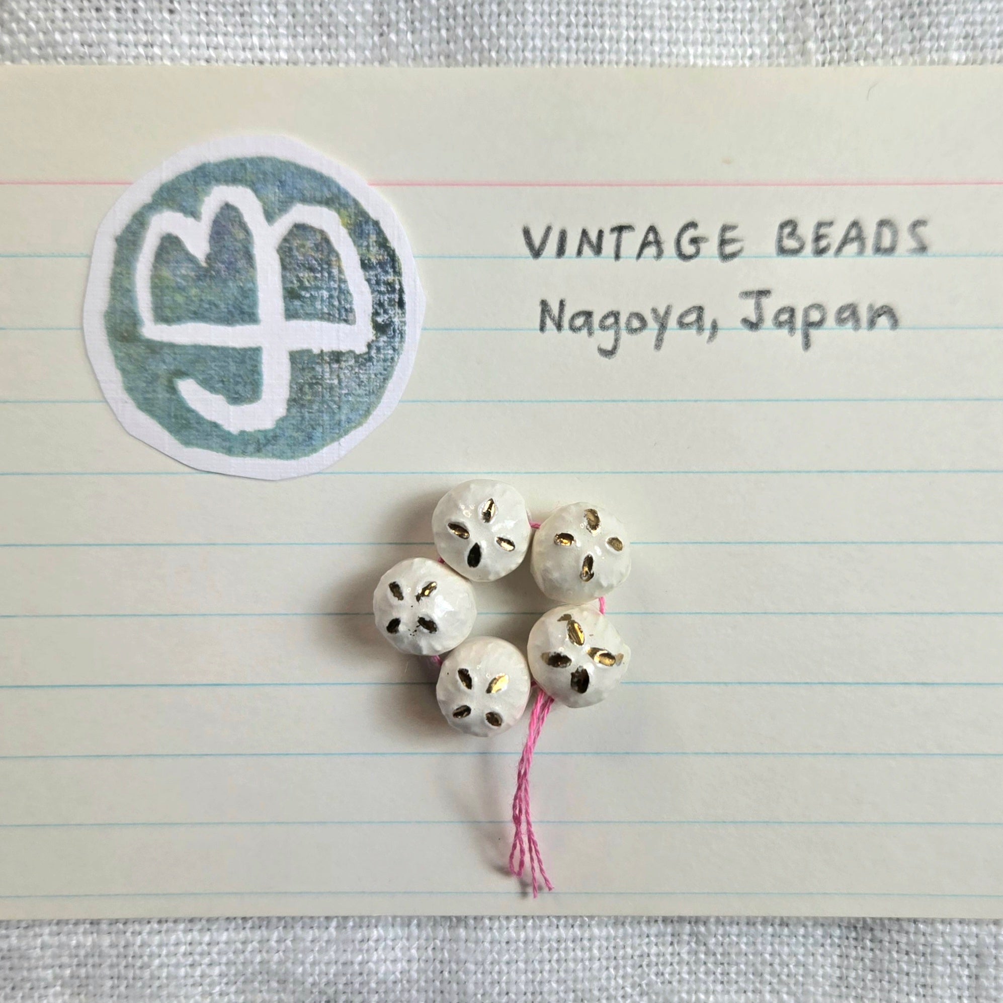 Five small vintage-style white beads with hand-painted black and gold details, tied to a pink string, displayed on a lined paper card with the text 'VINTAGE BEADS Nagoya, Japan' and a circular logo.