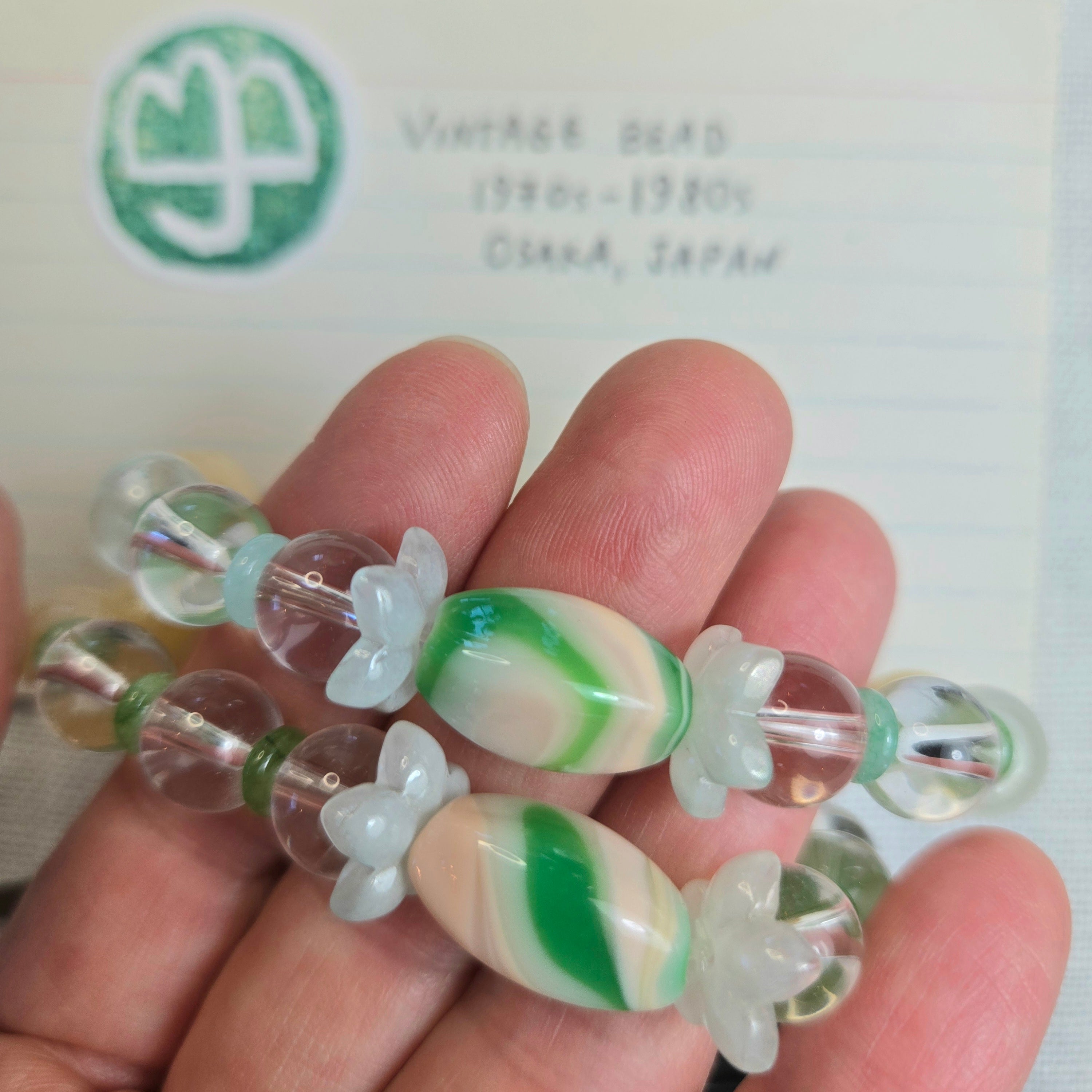 A close-up of a hand holding vintage Japanese bead jewelry, featuring translucent clear beads and two prominent green and cream marbled oval beads with white flower-shaped spacers. A handwritten note in the background reads 'VINTAGE BEAD 1980s-1980s OSAKA, JAPAN'.